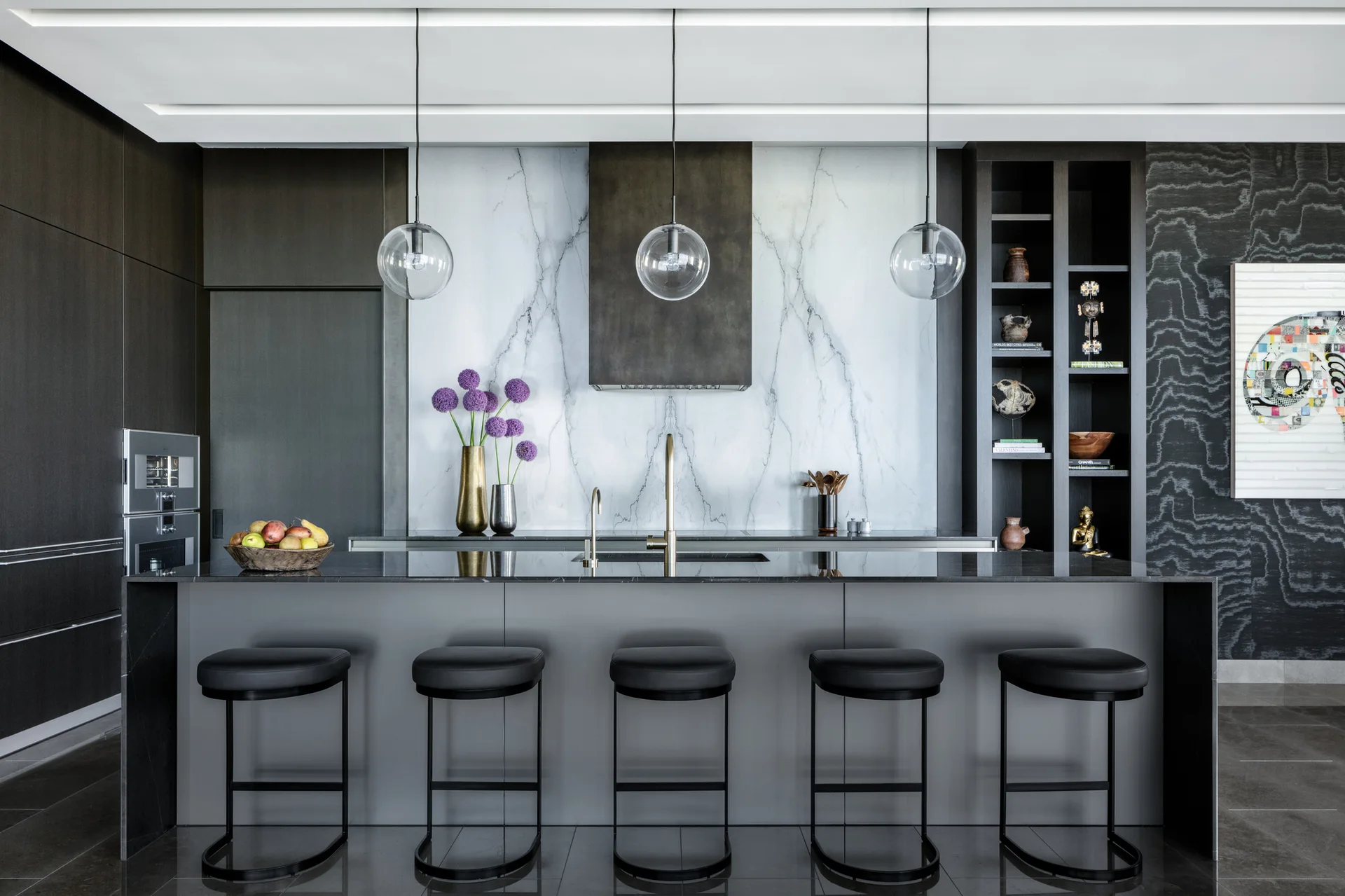 A dark gray kitchen island with five black bar stools sits in front of a white veined marble backsplash and glass globe pendant lights.