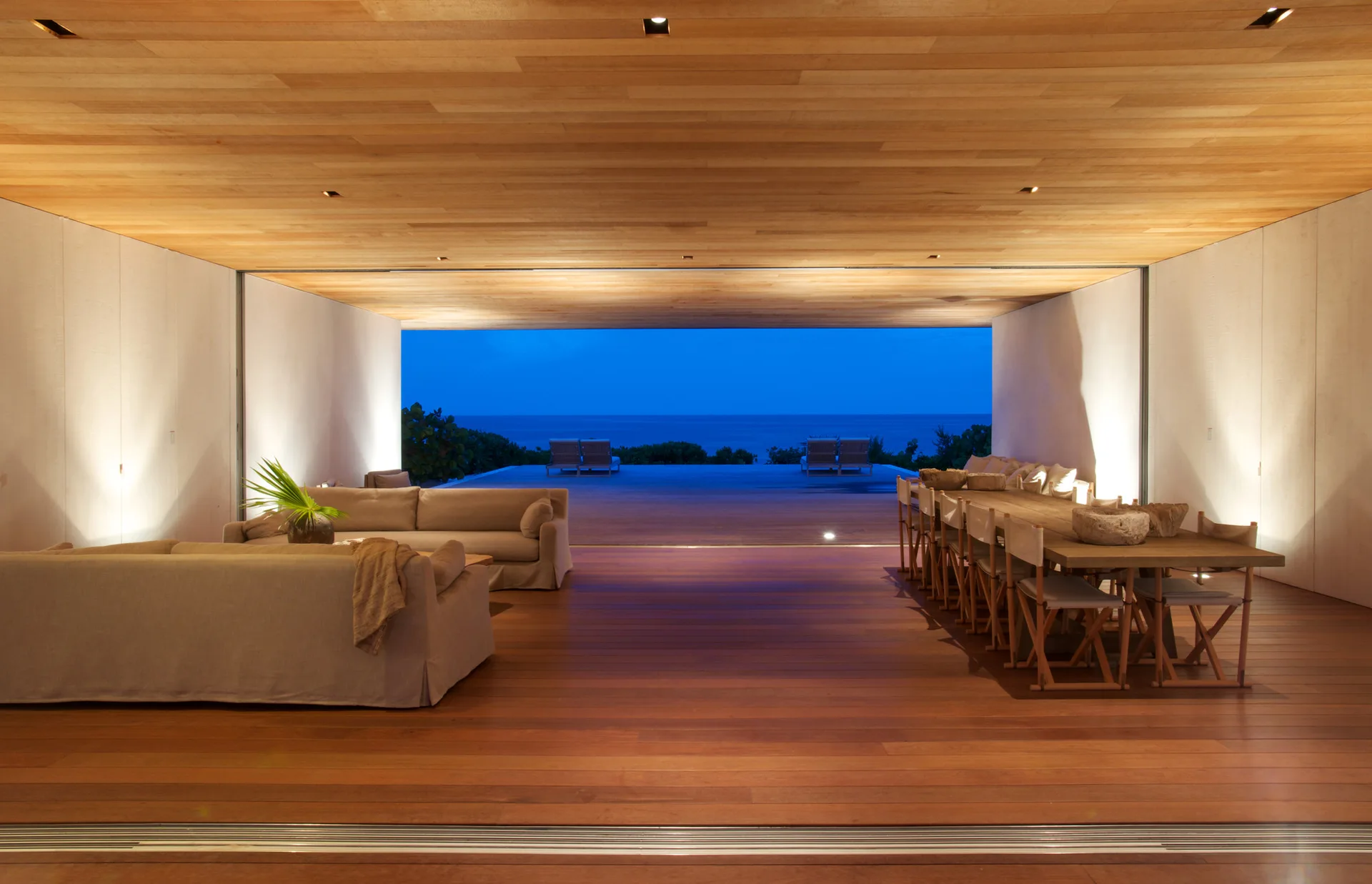An open-concept living and dining area with wood-paneled surfaces features neutral sofas, a long dining table, and an unobstructed view of the ocean.