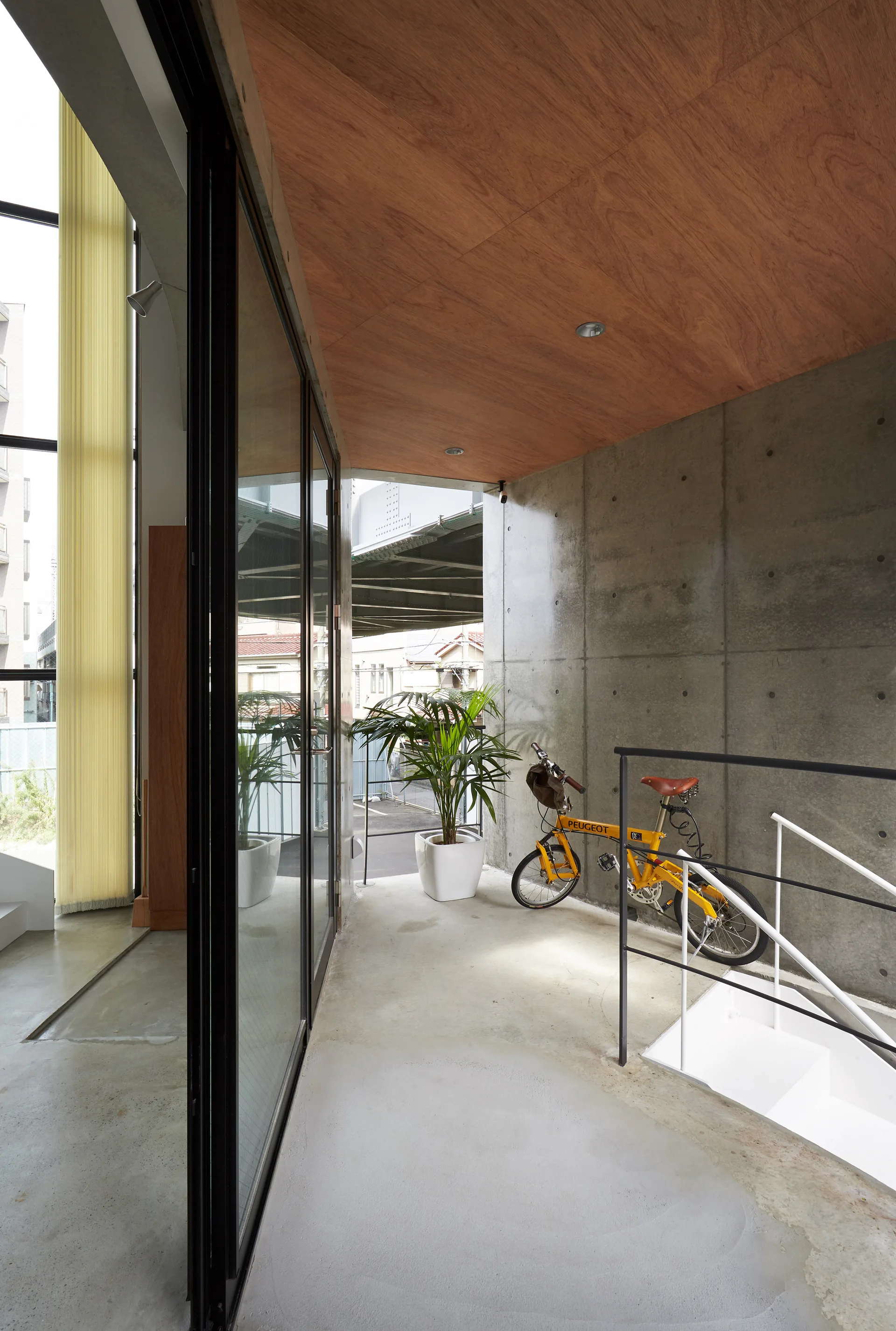 An industrial landing features a grey concrete wall, a warm wood paneled ceiling, a white staircase, and a yellow bicycle parked near a glass door.