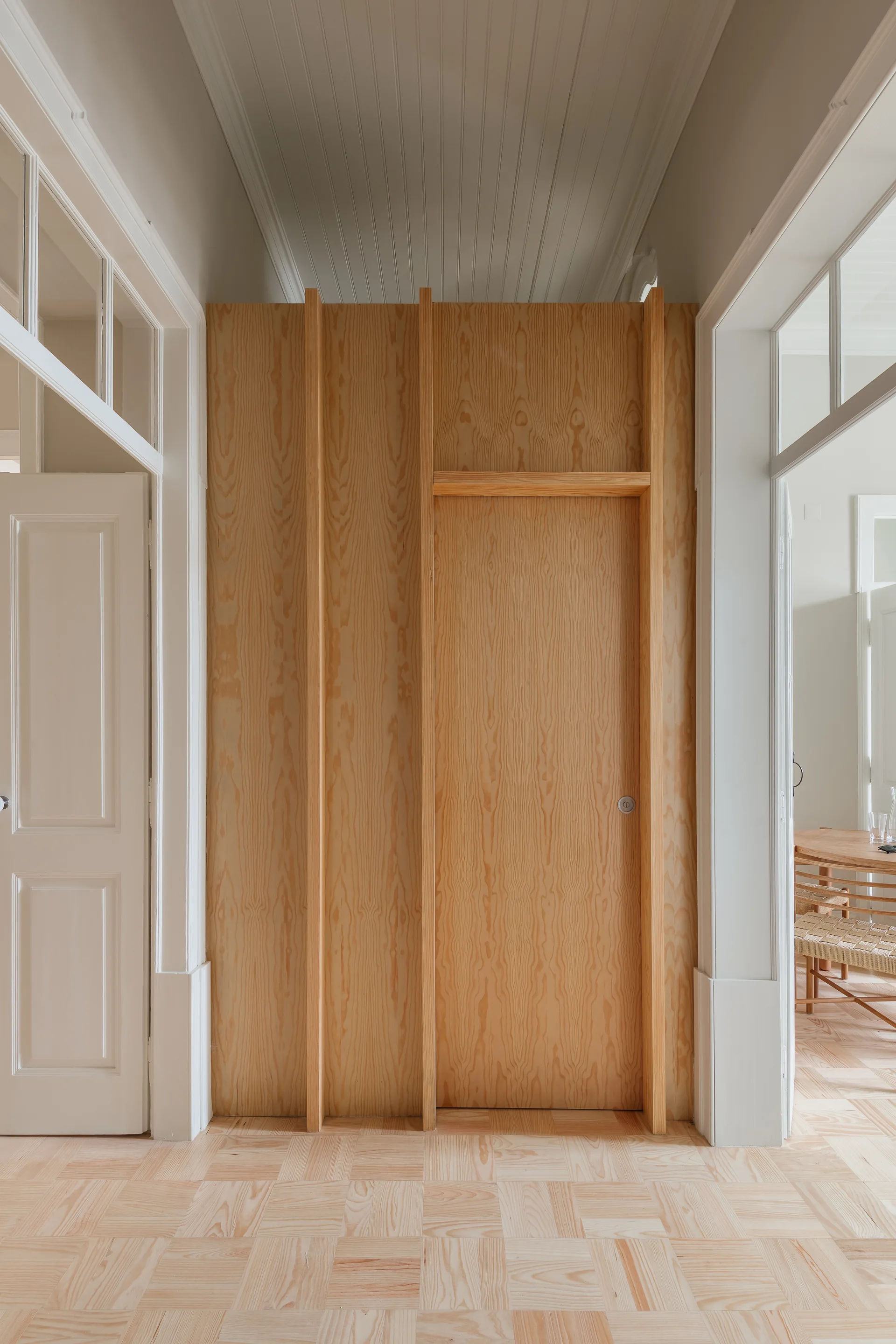A bright hallway features a light wood-paneled wall with a flush integrated door, parquet flooring, white trim, and a high ceiling.