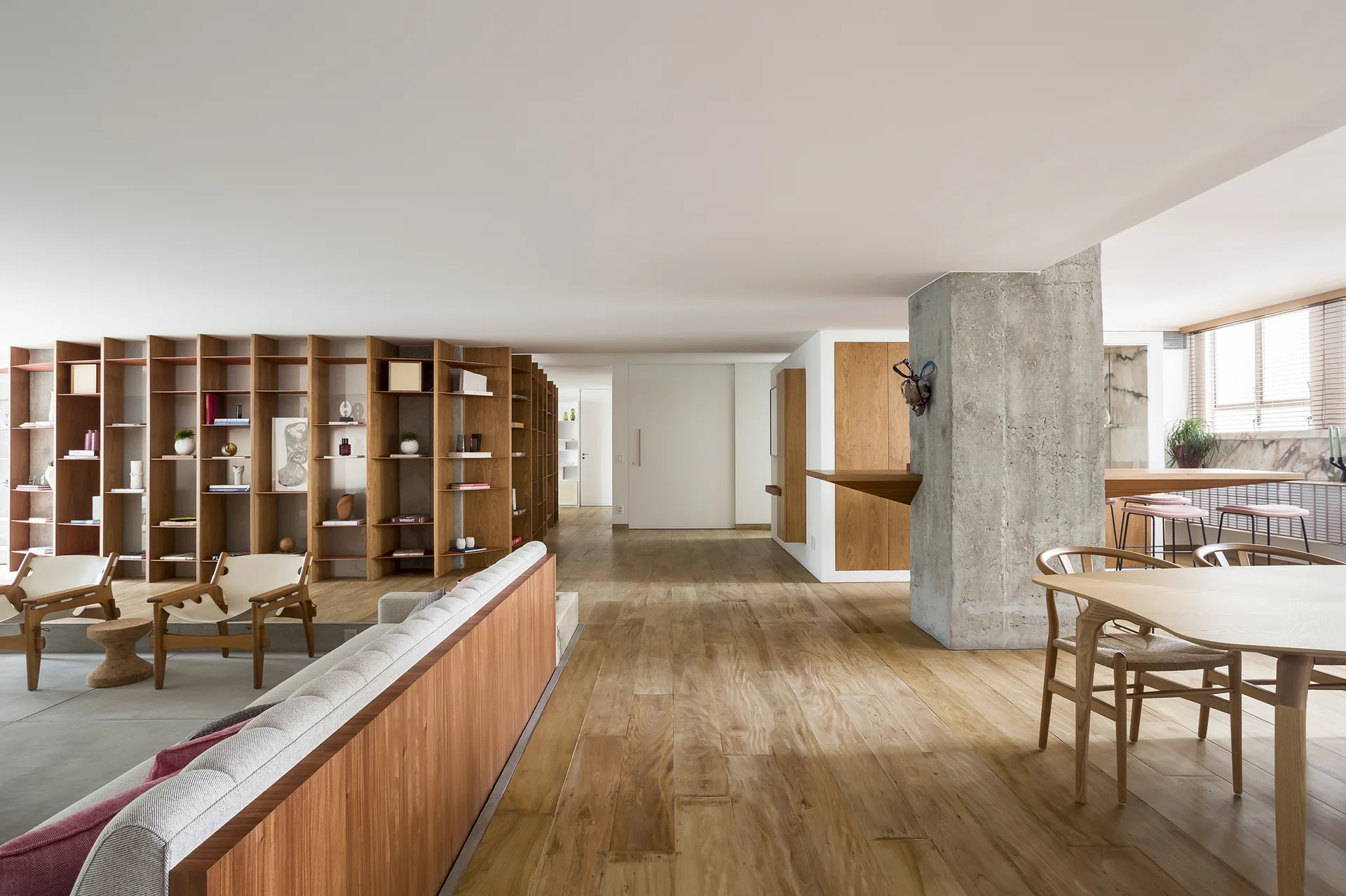An open-plan living room with a large wooden bookshelf partition, an exposed concrete pillar, wood floors, and a dining table.