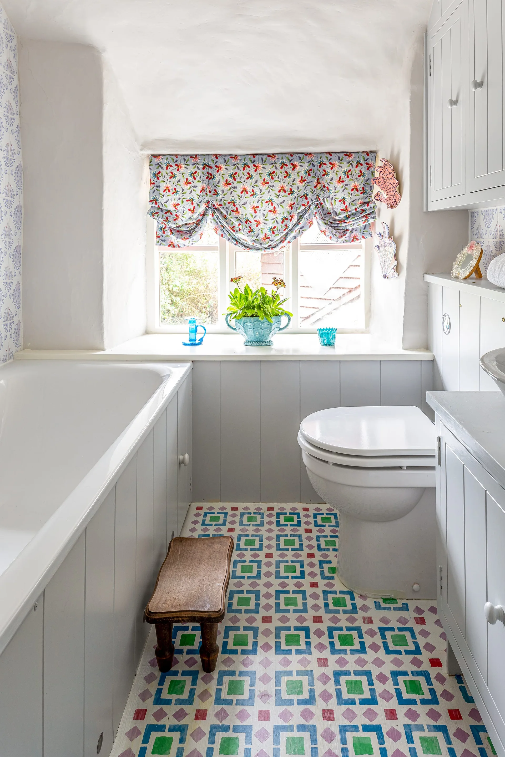 A small cottage bathroom features a white bathtub, toilet, gray vertical wood paneling, and a colorful geometric patterned floor under a window.