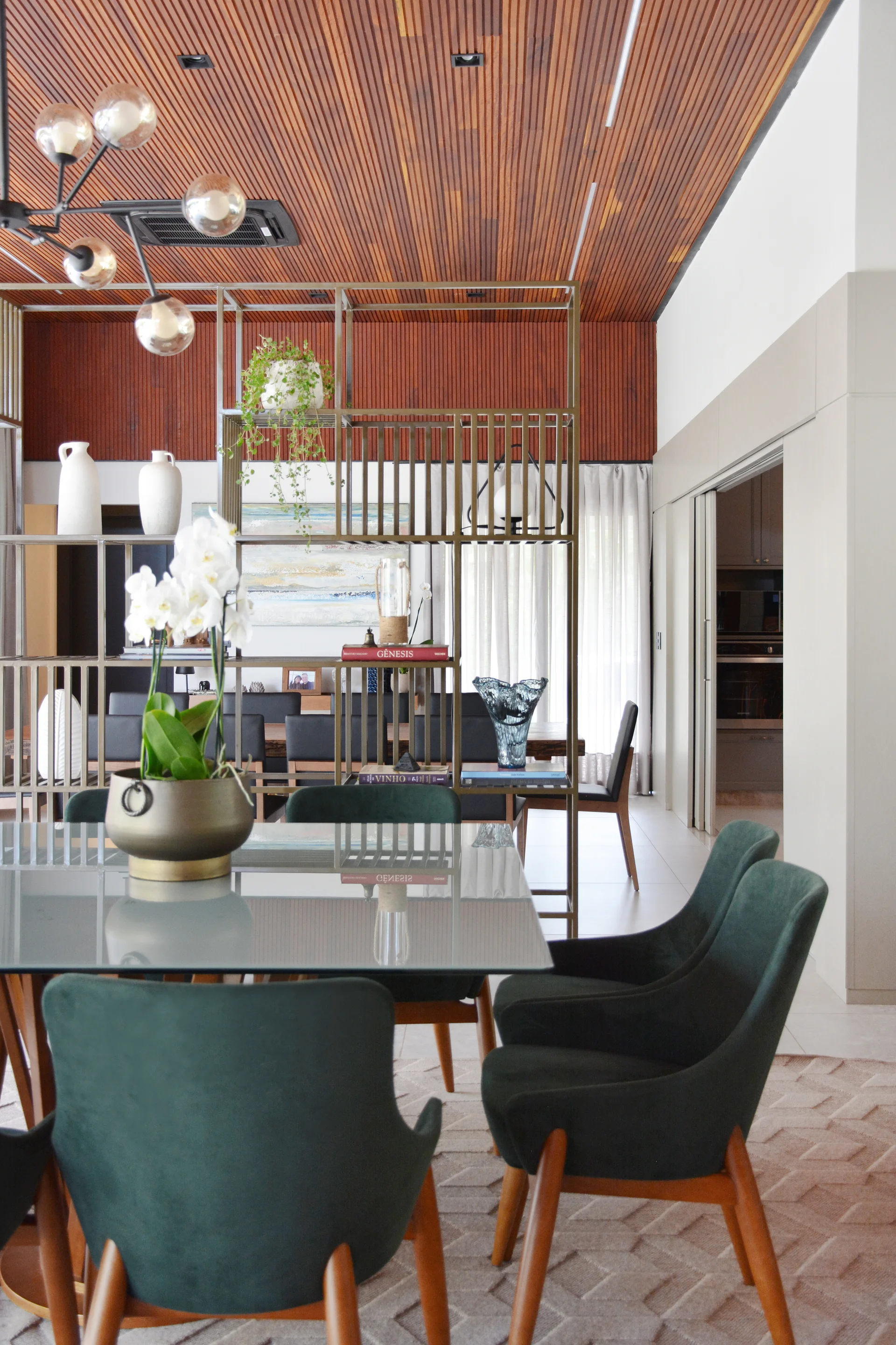 A contemporary dining room features dark green upholstered chairs, a glass-top table, a wood slat ceiling, and an open metal shelving divider.