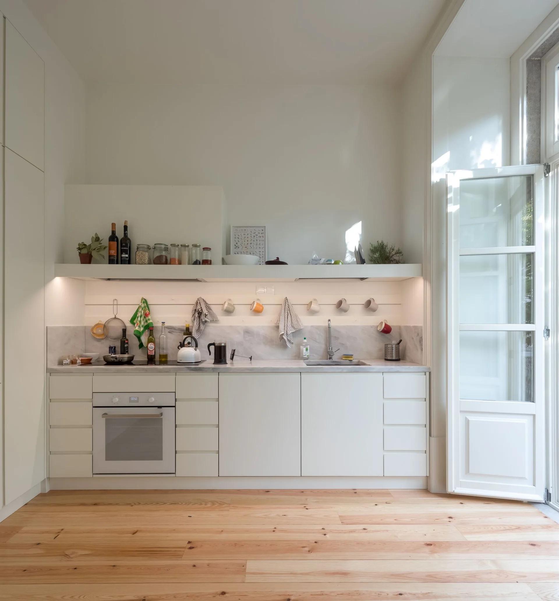 A minimalist white kitchen features flat-front cabinets, a marble-like countertop and backsplash, and a long floating shelf with under-cabinet lighting.