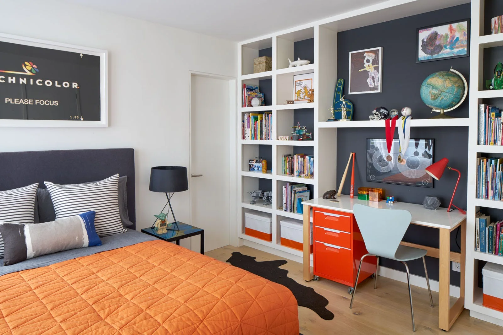A modern kids bedroom featuring a bed with an orange quilt, a white built-in bookcase with a desk nook, and a charcoal gray accent wall.