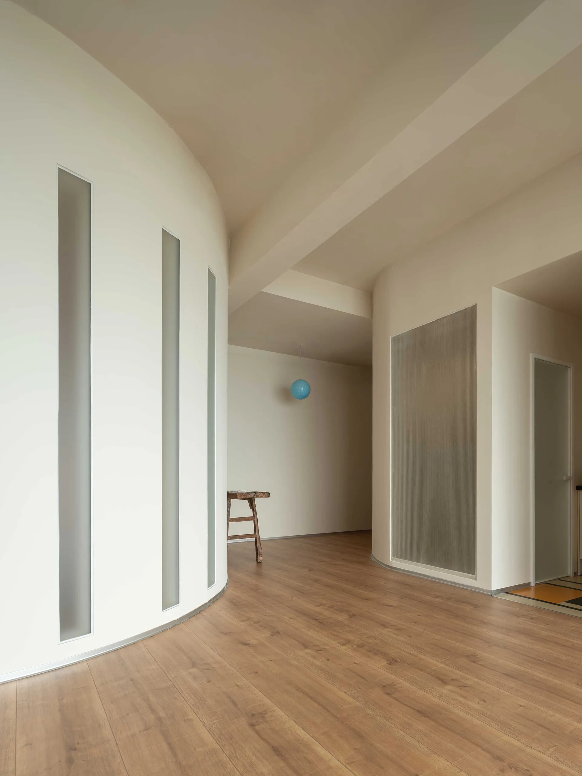 A minimalist hallway features a curved white wall with three tall, thin windows, wood flooring, and a ribbed glass partition.