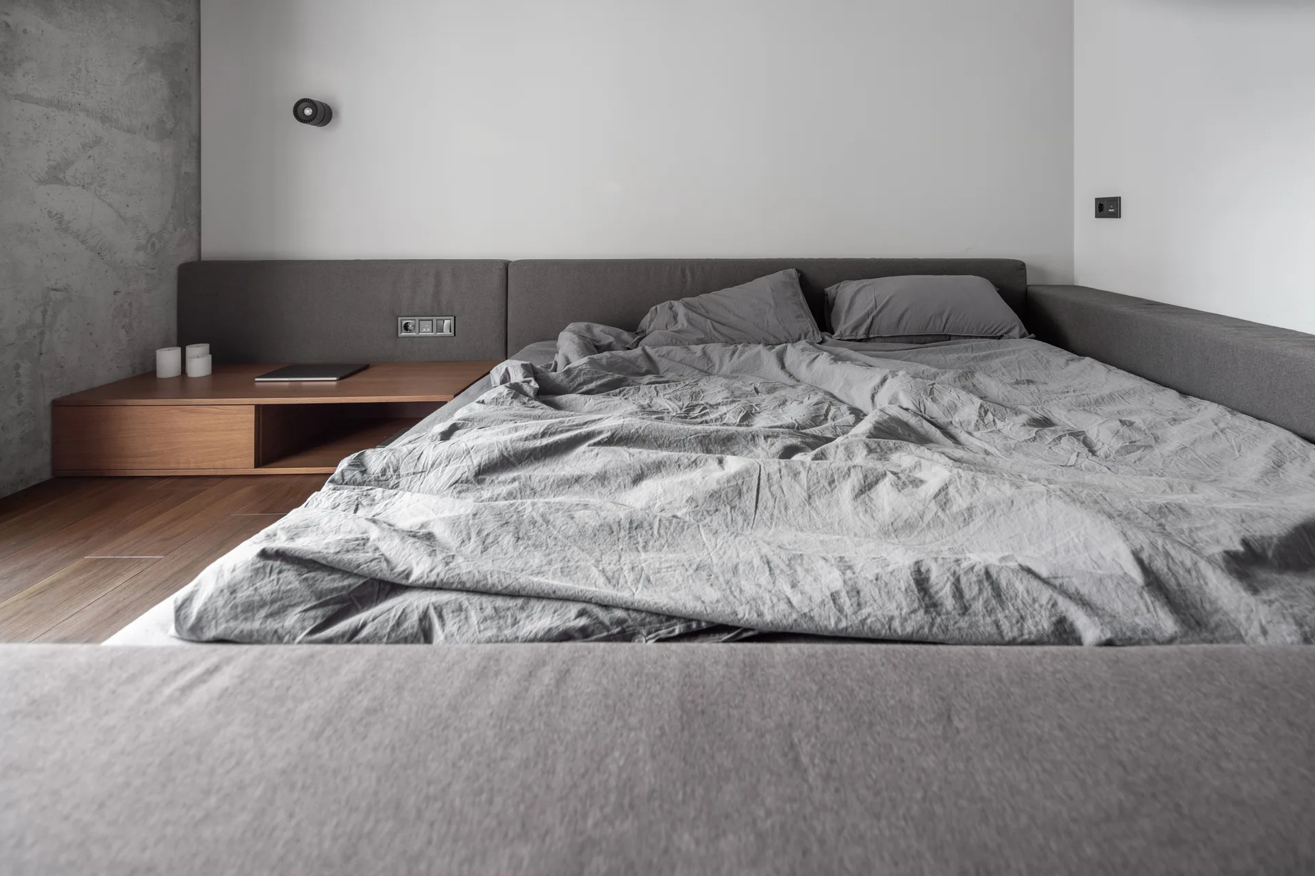 A low grey upholstered bed with rumpled linen bedding is paired with an integrated wooden bedside platform against white and concrete walls.