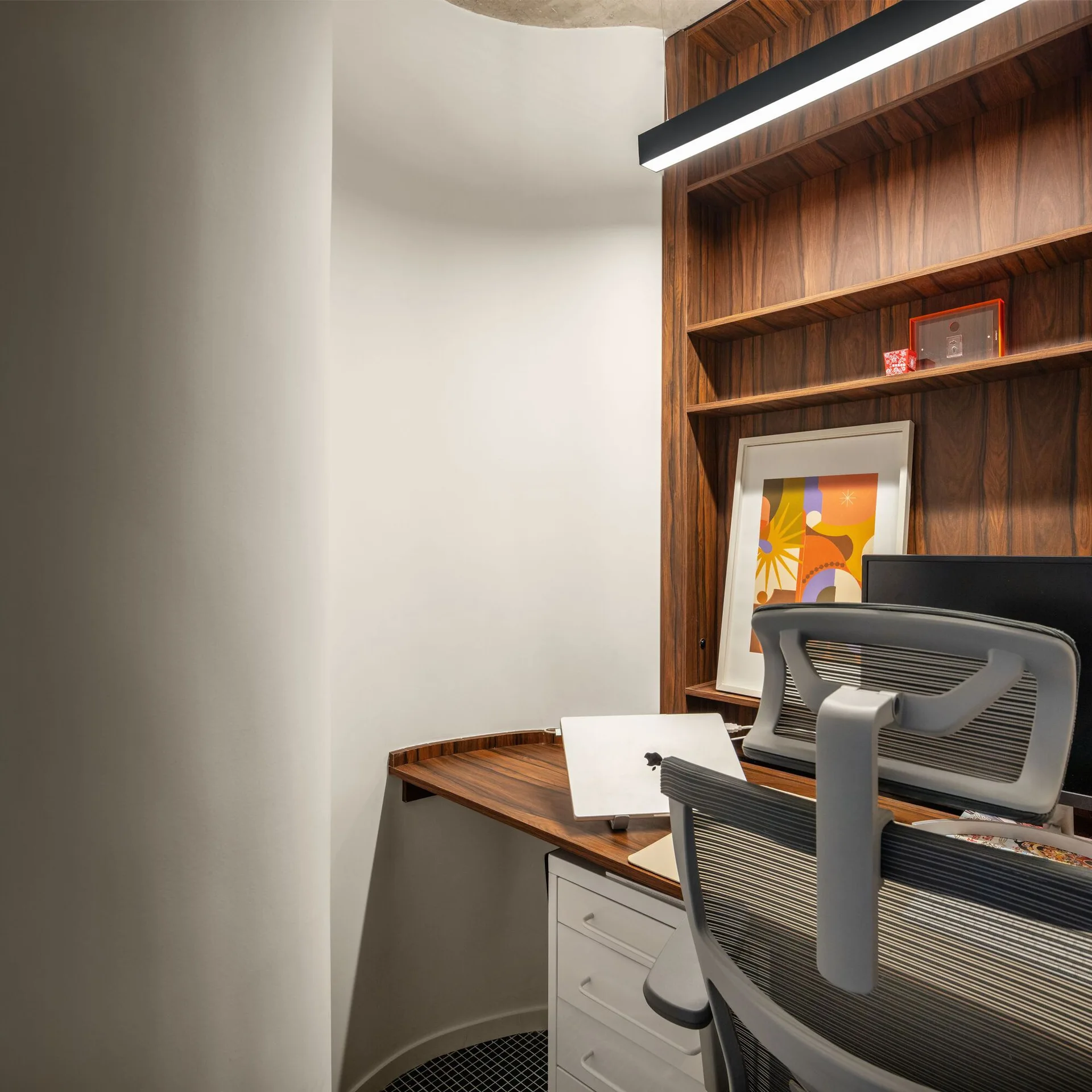 A home office corner featuring a built-in wood desk and shelving unit, a grey ergonomic chair, and a smooth white curved wall.