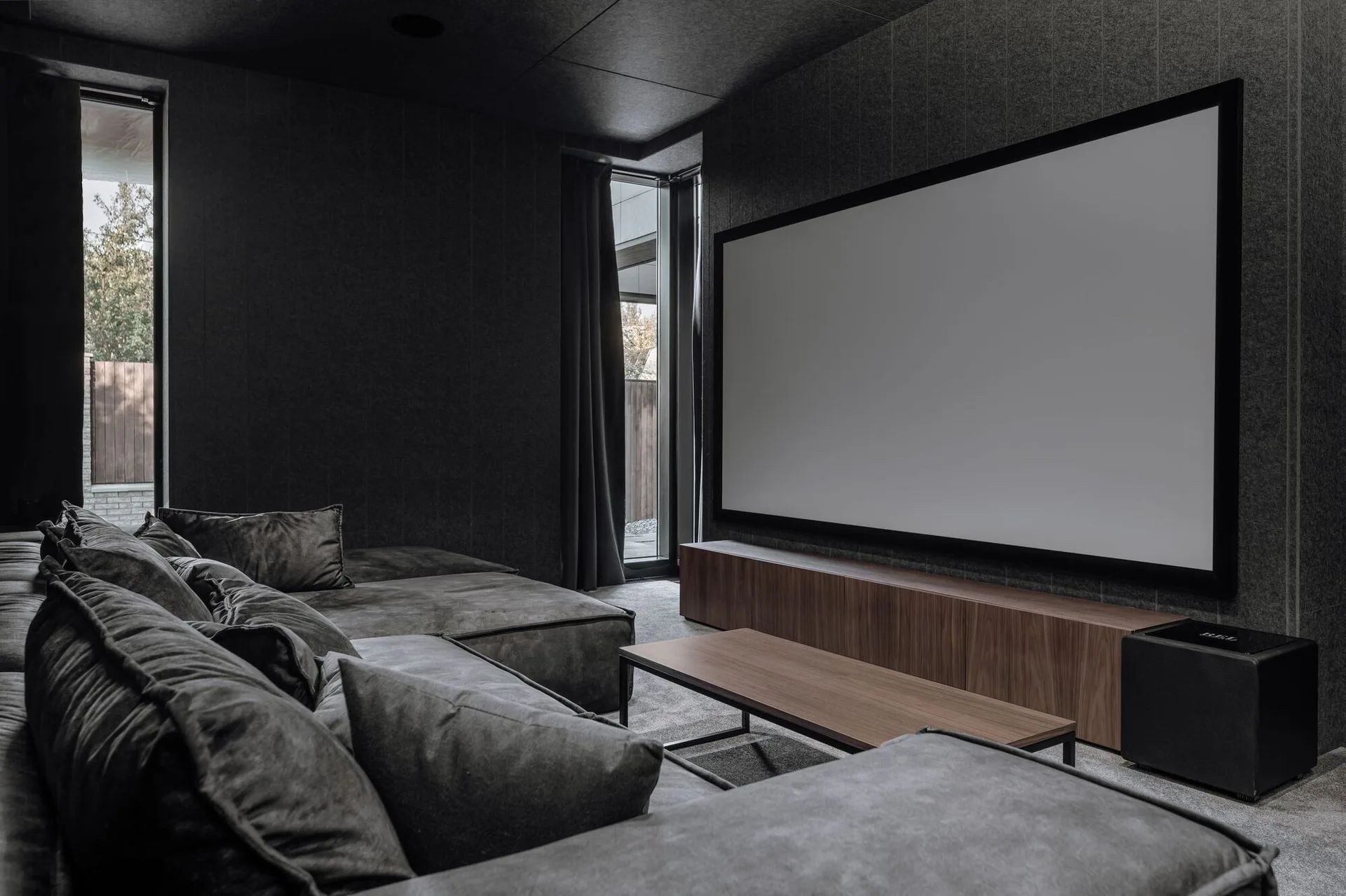 A dimly lit home theater features a large wall-mounted projection screen above a low wooden console and a plush grey sectional sofa.