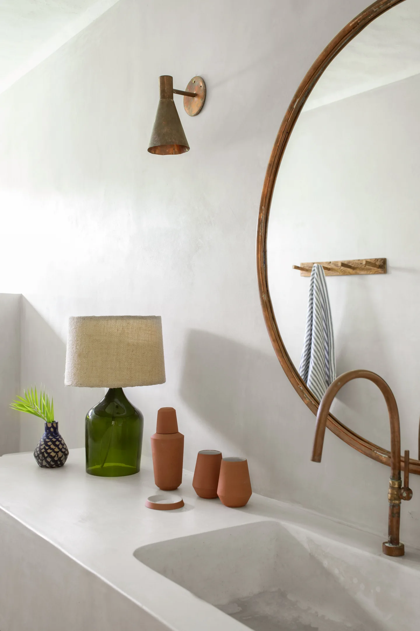 A plaster bathroom vanity features an integrated sink, a round copper mirror, a green glass table lamp, and terracotta accessories.