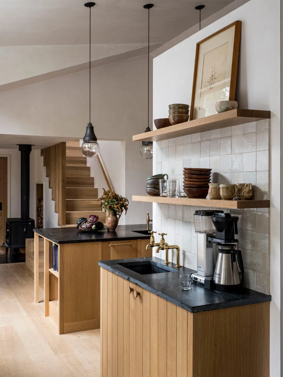 A kitchen featuring oak cabinetry under a dark stone countertop with a brass faucet, topped by floating wood shelves against a square tiled backsplash.