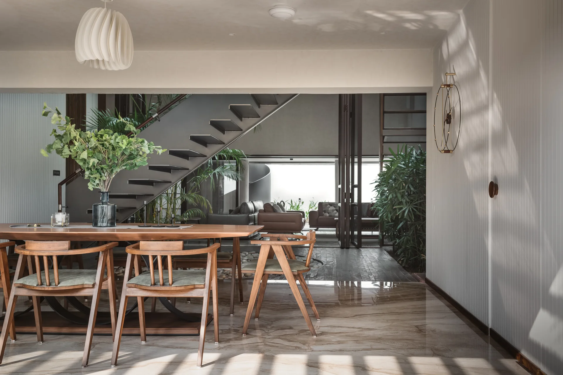 A wooden dining table and chairs sit on a polished marble floor next to a white grooved wall with an open-riser staircase in the background.