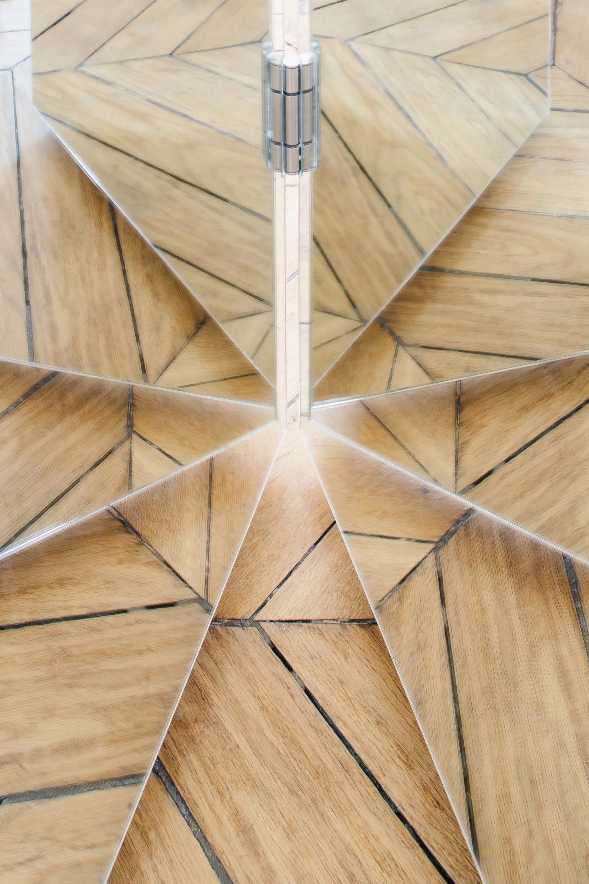 A close-up view of light brown geometric wood floor planks reflected in multiple angled mirror panels connected by a silver hinge.