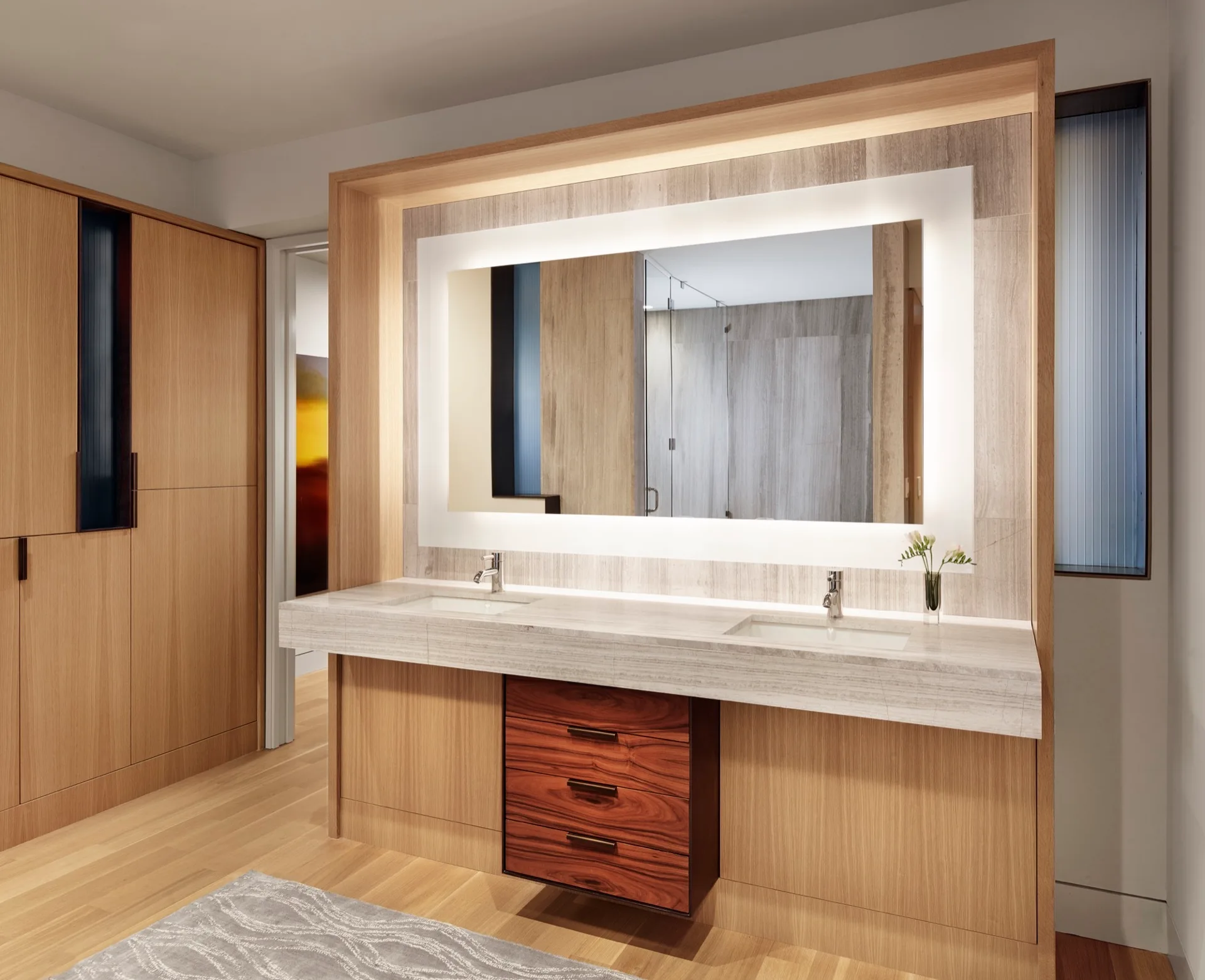 A modern bathroom features a double wood vanity with a stone countertop, backlit rectangular mirror, and a contrasting central floating drawer unit.