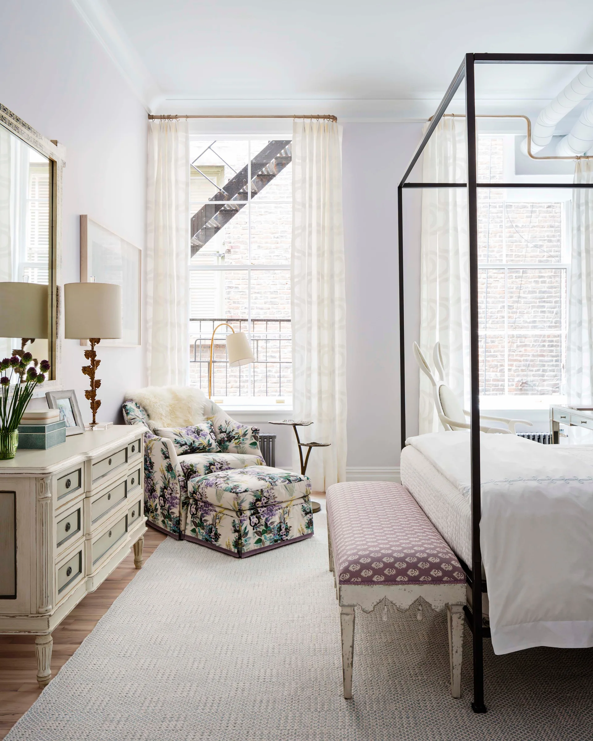 A bright bedroom featuring a black metal canopy bed, a floral upholstered armchair with matching ottoman, and a distressed white wooden dresser.