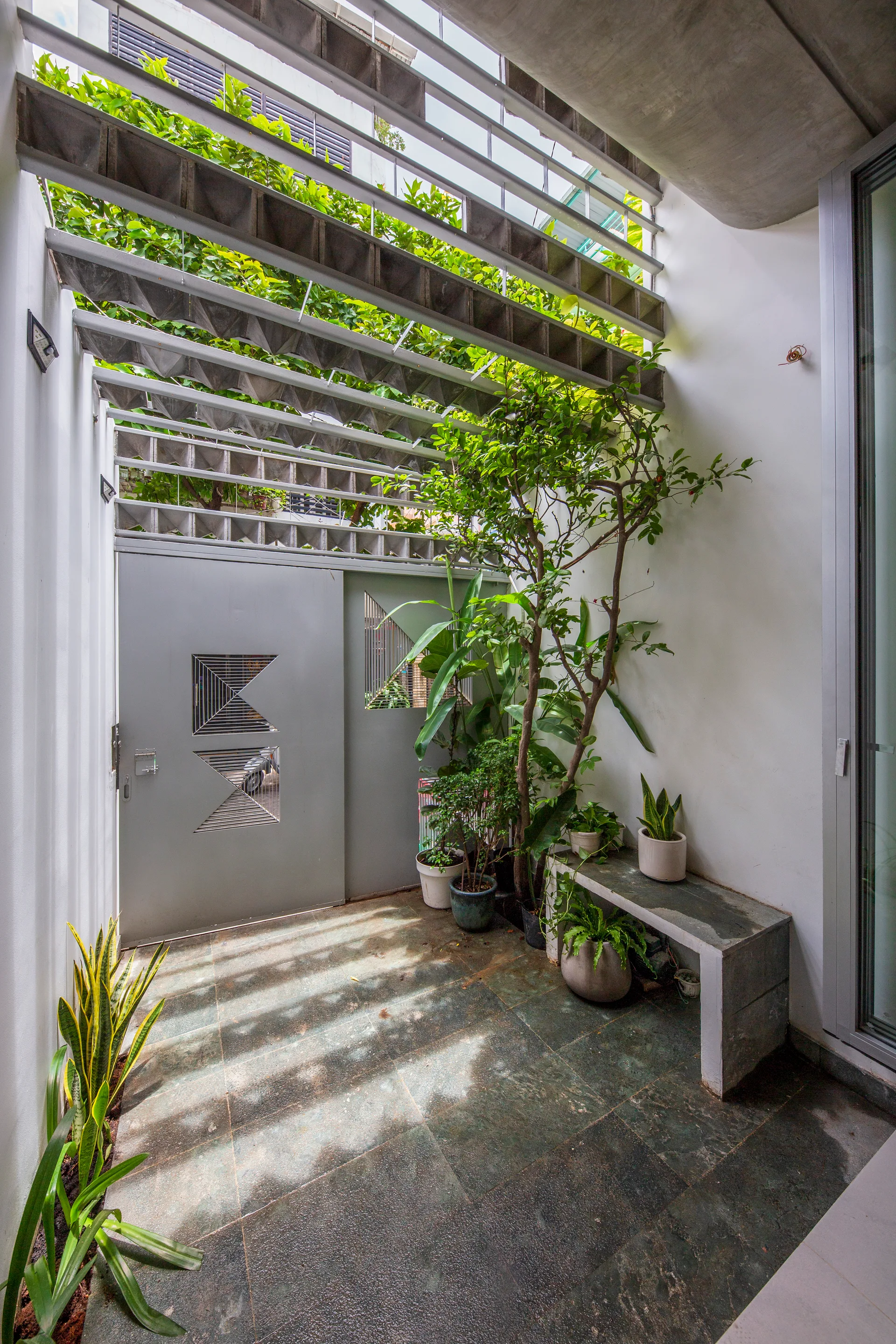 A modern outdoor courtyard featuring a grey metal gate with geometric cutouts, a slatted concrete pergola overhead, and potted plants.