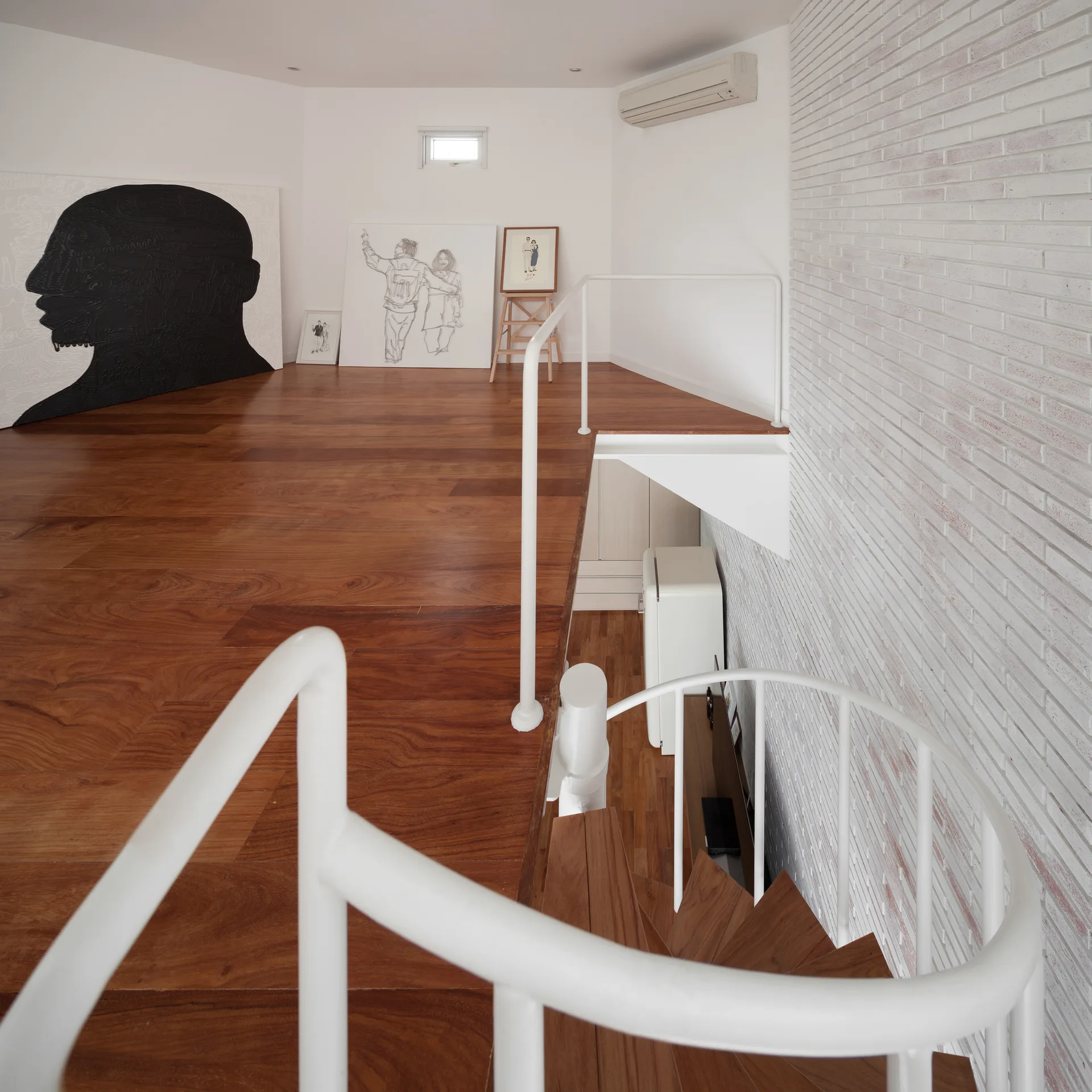 A white metal spiral staircase leads to a wooden-floored mezzanine level beside a textured white painted brick wall.