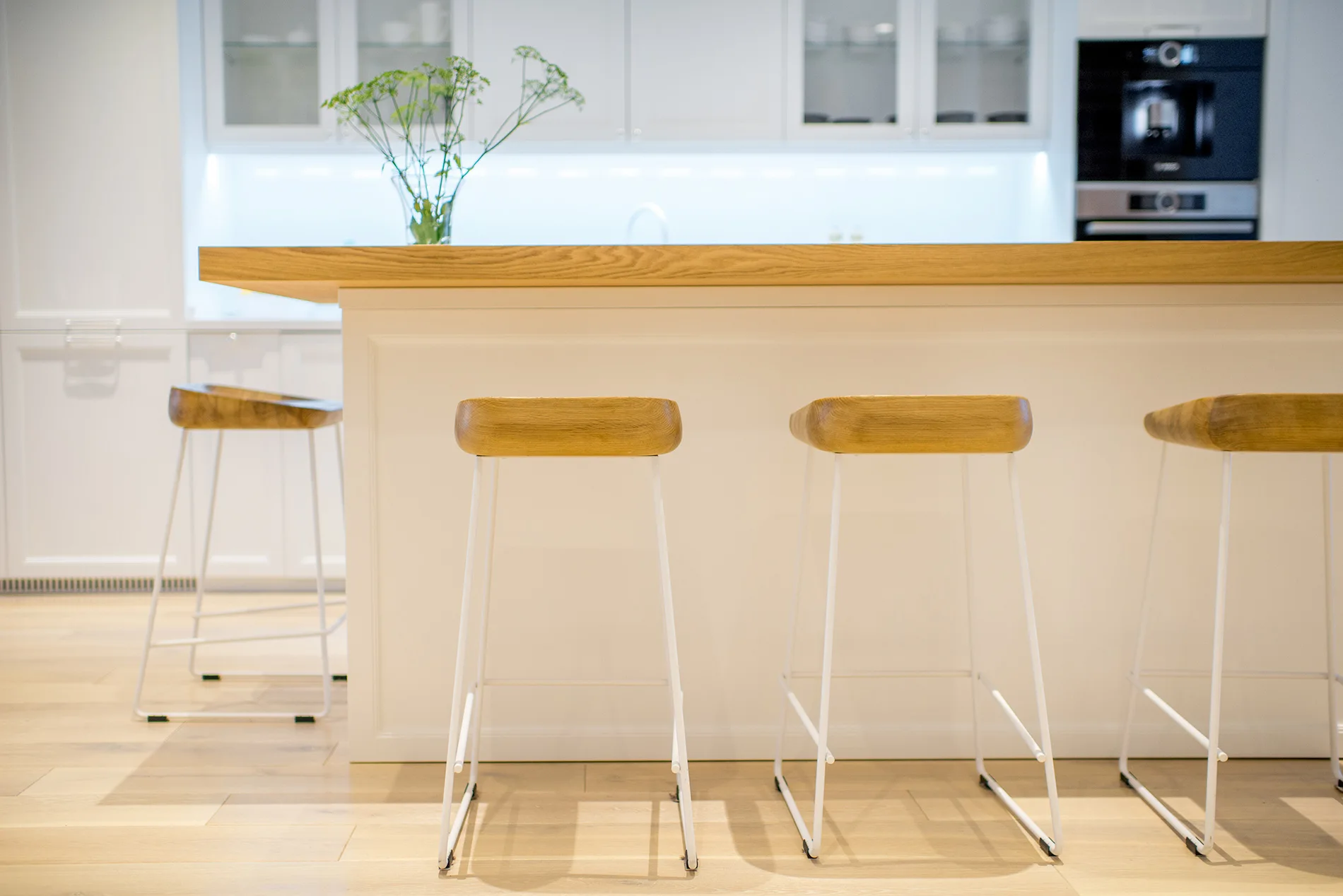 Three minimalist bar stools with white metal legs and wooden seats stand next to a white kitchen island with a thick wood countertop.
