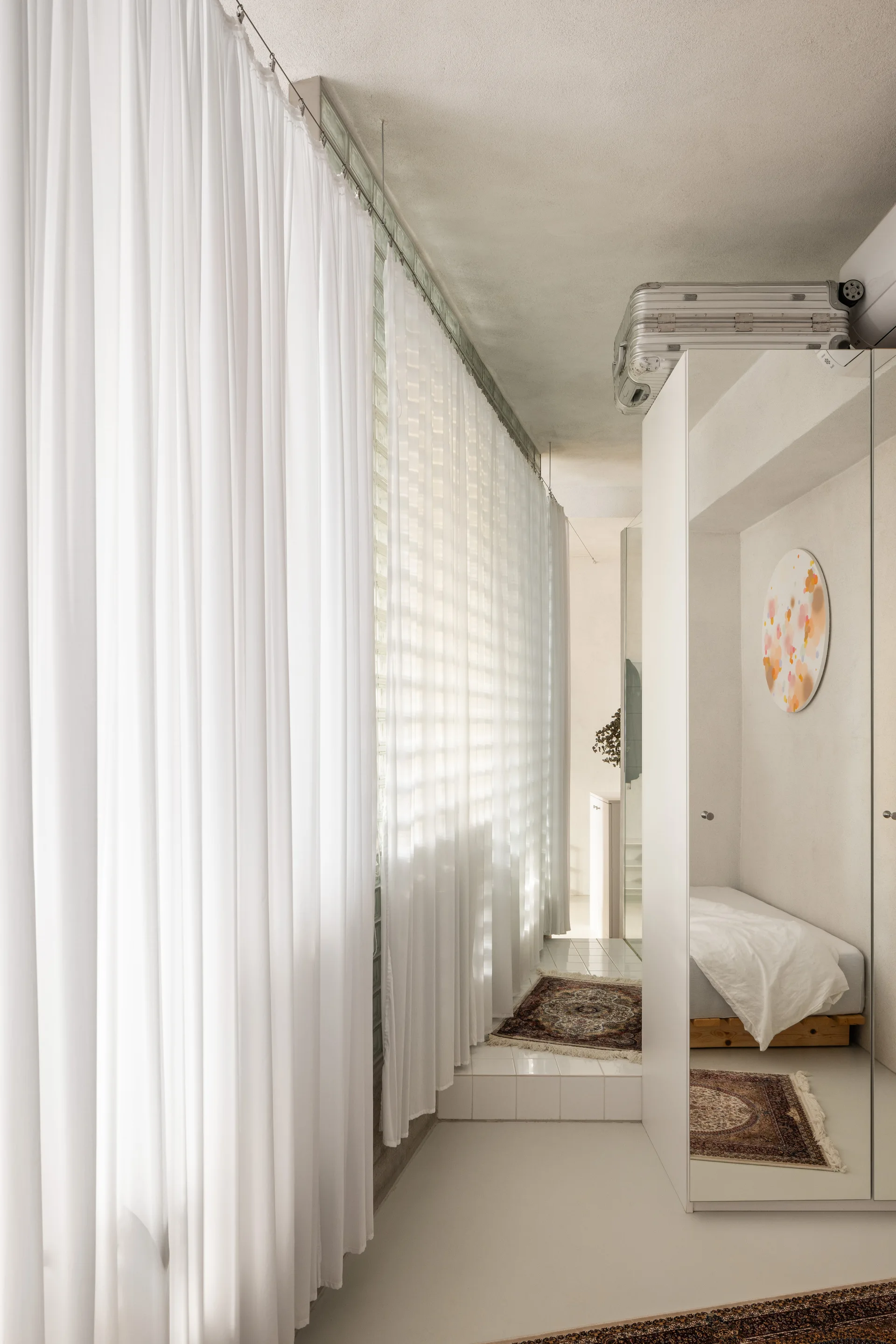 A minimalist white bedroom featuring a glass block wall with sheer curtains and full-length mirrored closet doors.