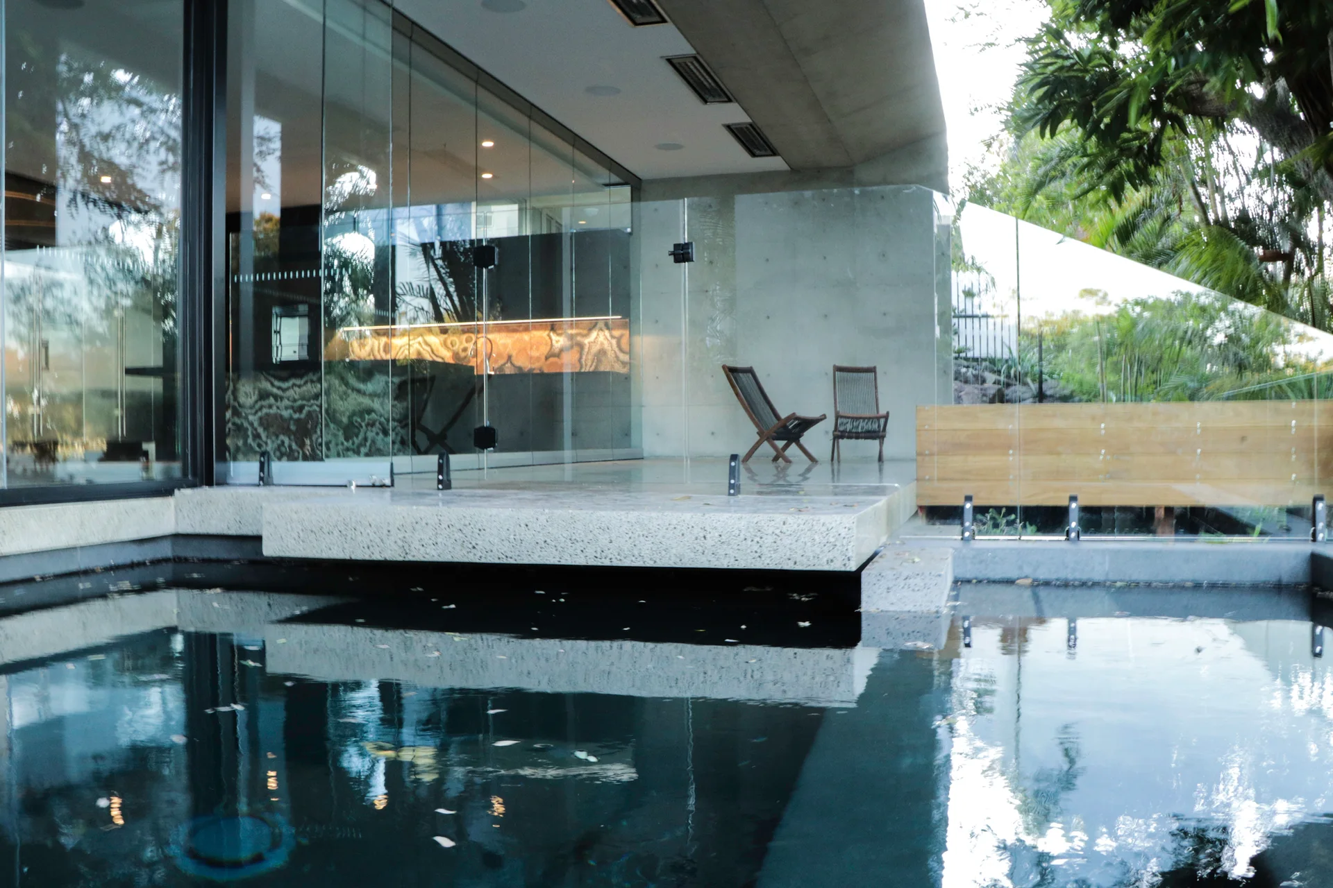 A modern concrete patio features a dark swimming pool in the foreground, frameless glass walls, and two wooden folding chairs.