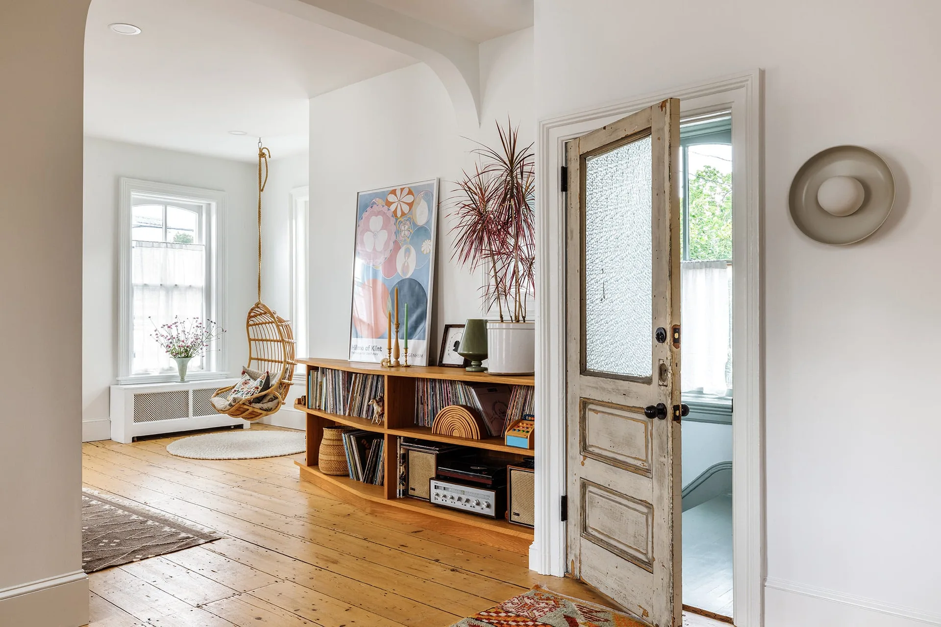 A bright living room with light wood floors, a low wooden bookshelf filled with books, a hanging rattan chair, and a distressed vintage door.