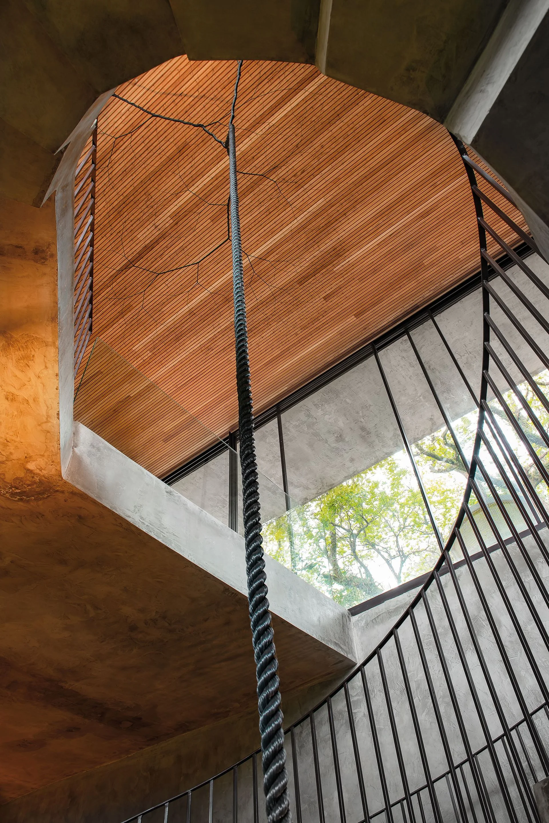 A thick black rope hangs vertically down the center of an open staircase with concrete walls and a slatted wood ceiling.