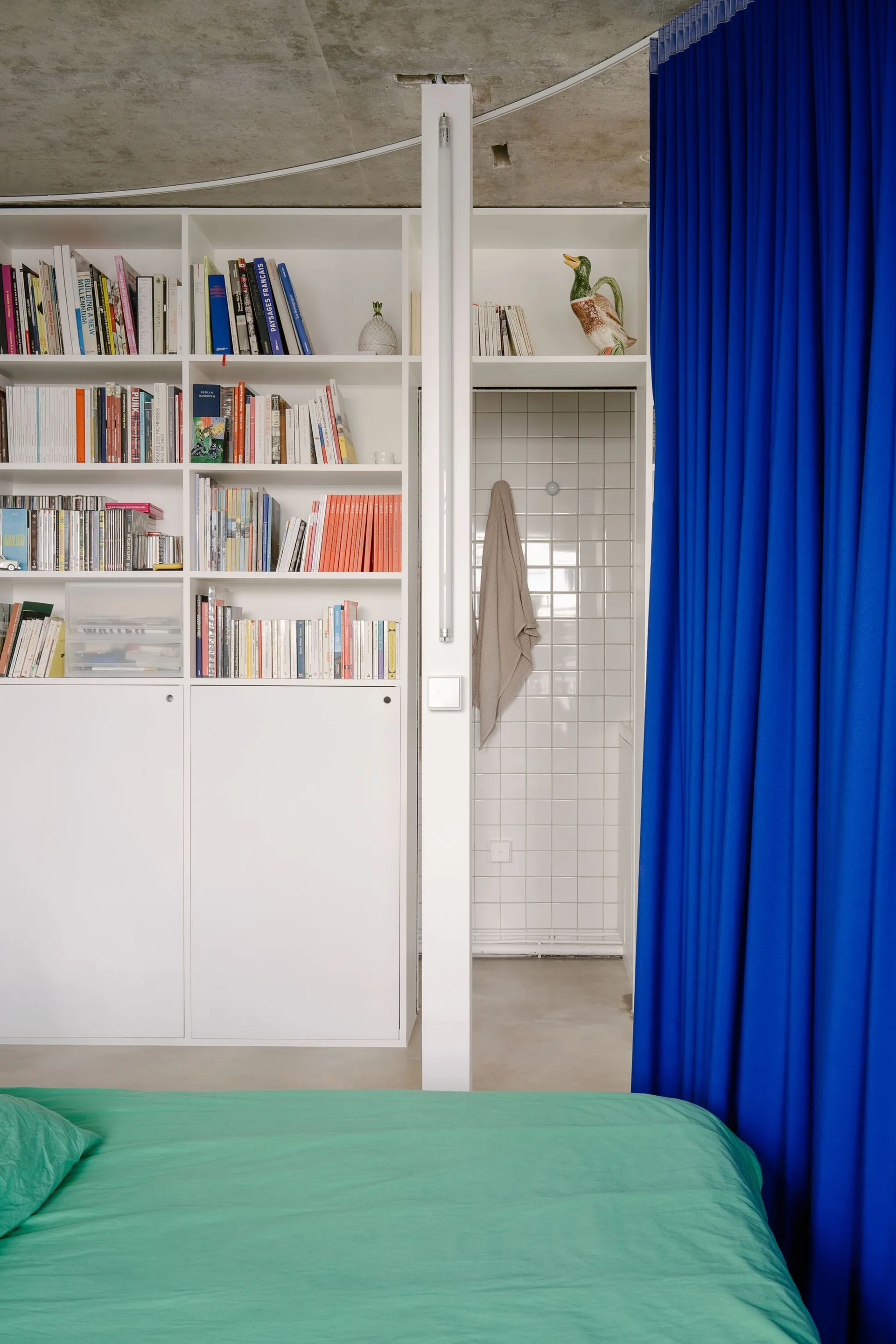 A small studio bedroom featuring a green bed in the foreground, a white built-in bookshelf, and a cobalt blue room-dividing curtain.