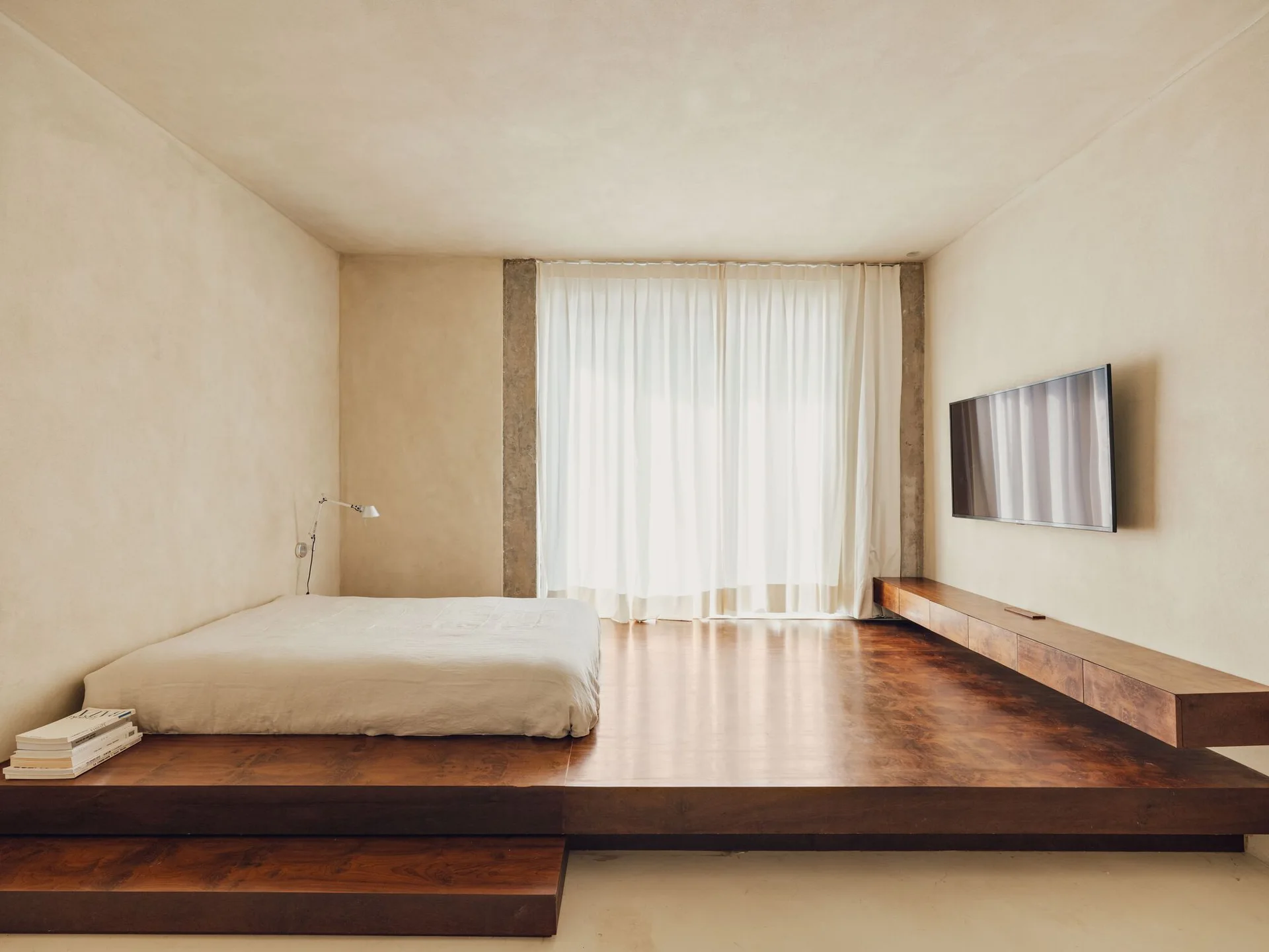 A minimalist bedroom features a low platform bed on a raised dark wood floor, warm beige plaster walls, a floating wood console, and a wall-mounted TV.