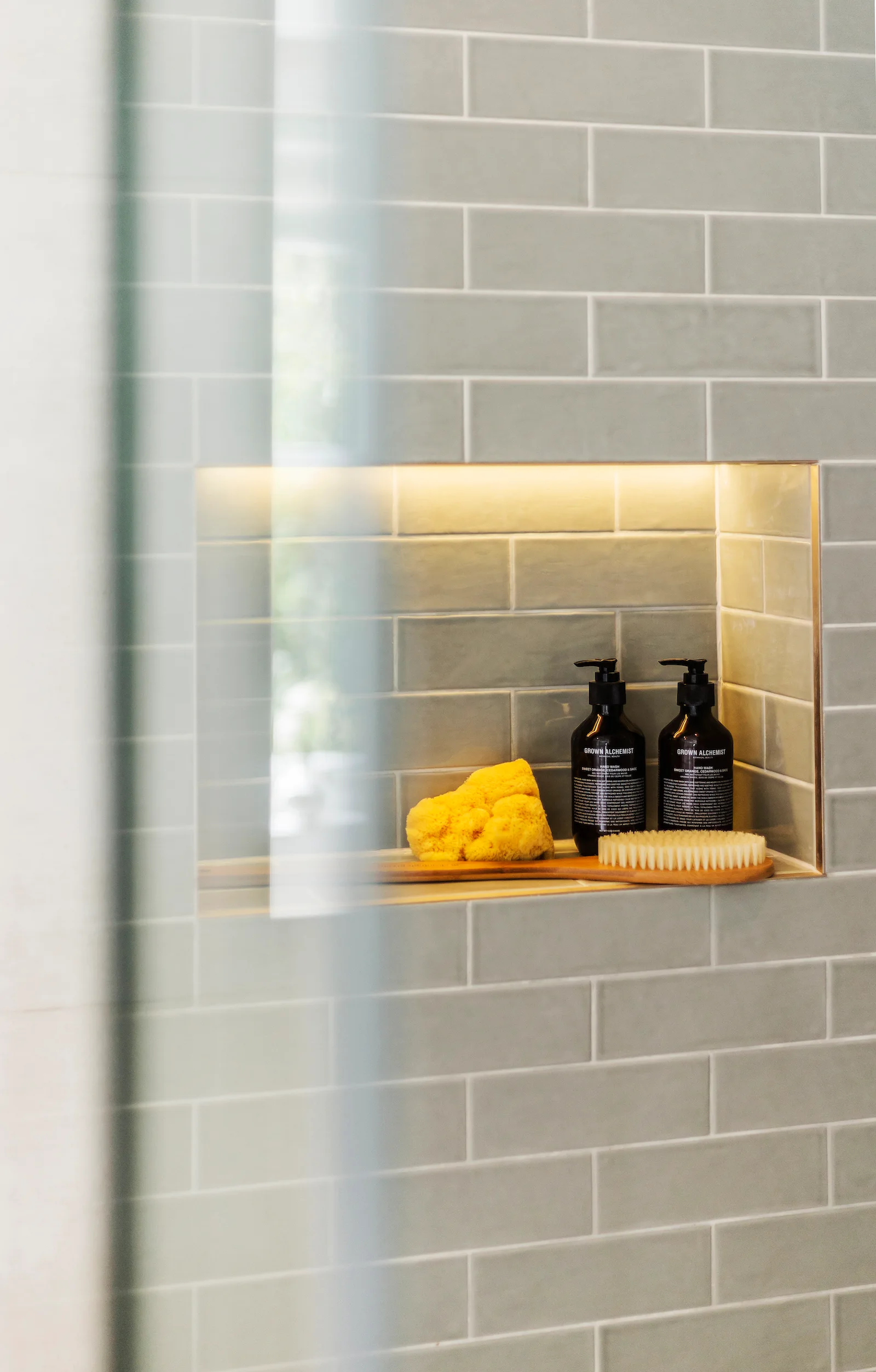 A recessed shower niche in a sage green subway tiled wall features integrated warm lighting, holding two black bottles, a wooden brush, and a yellow sponge.