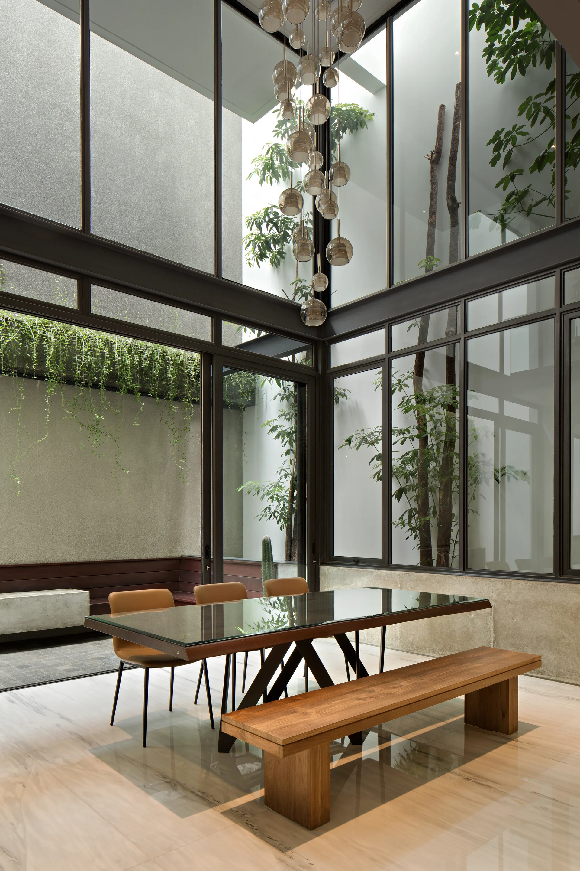 A modern dining room features a glass-top table, tan chairs, a wooden bench, and floor-to-ceiling glass walls looking out to a courtyard garden.