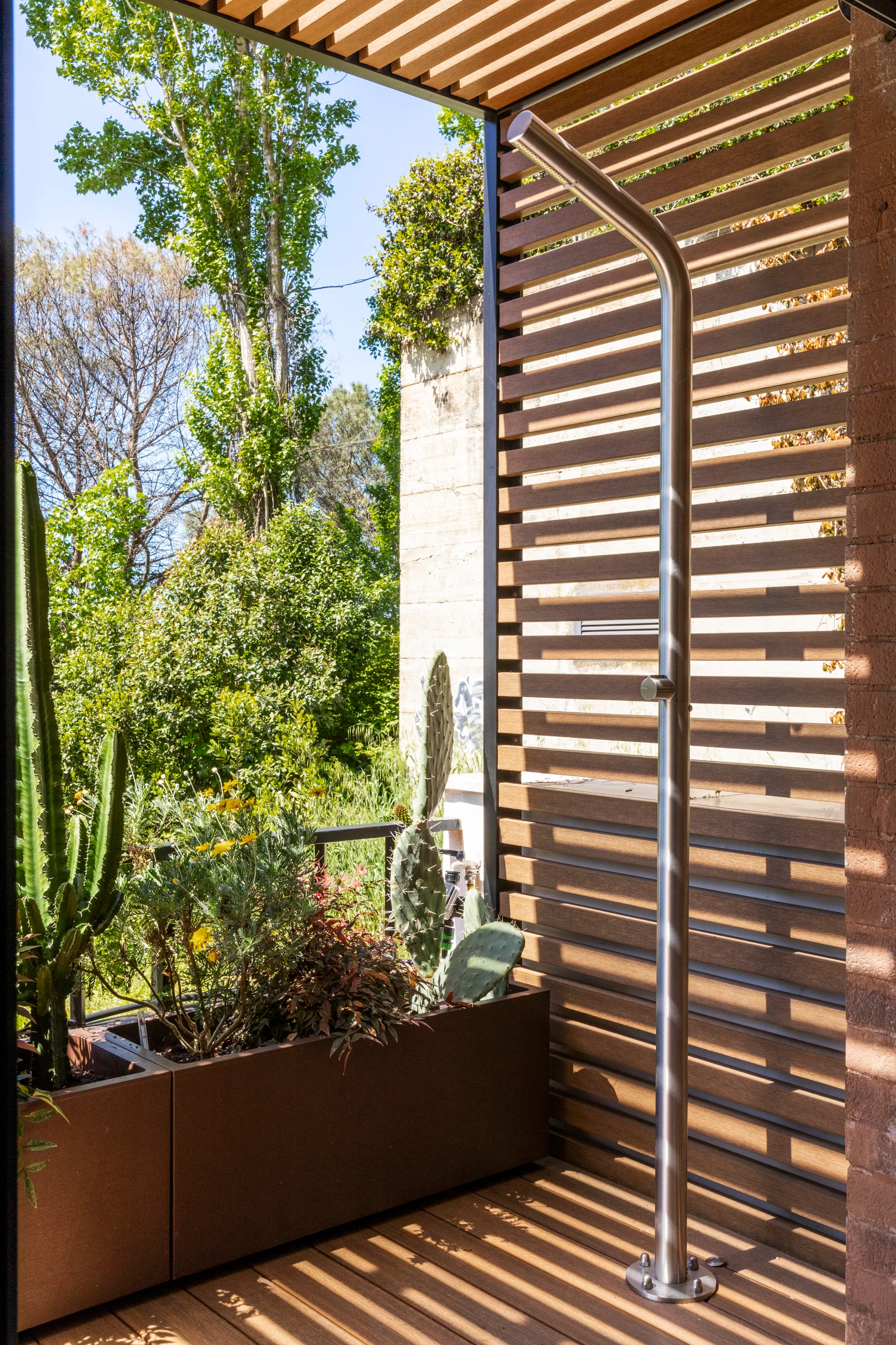 A modern metallic outdoor shower fixture mounted next to a horizontal slatted wooden privacy wall with a potted cactus planter.