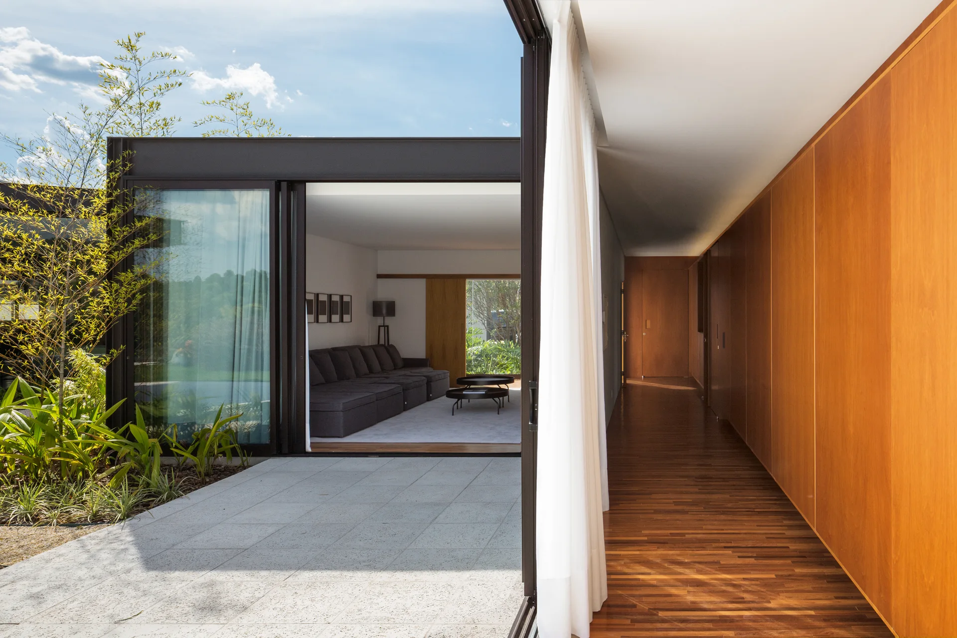 A modern living room with a grey sectional sofa connects to an outdoor patio next to a long hallway lined with warm wood wall panels.
