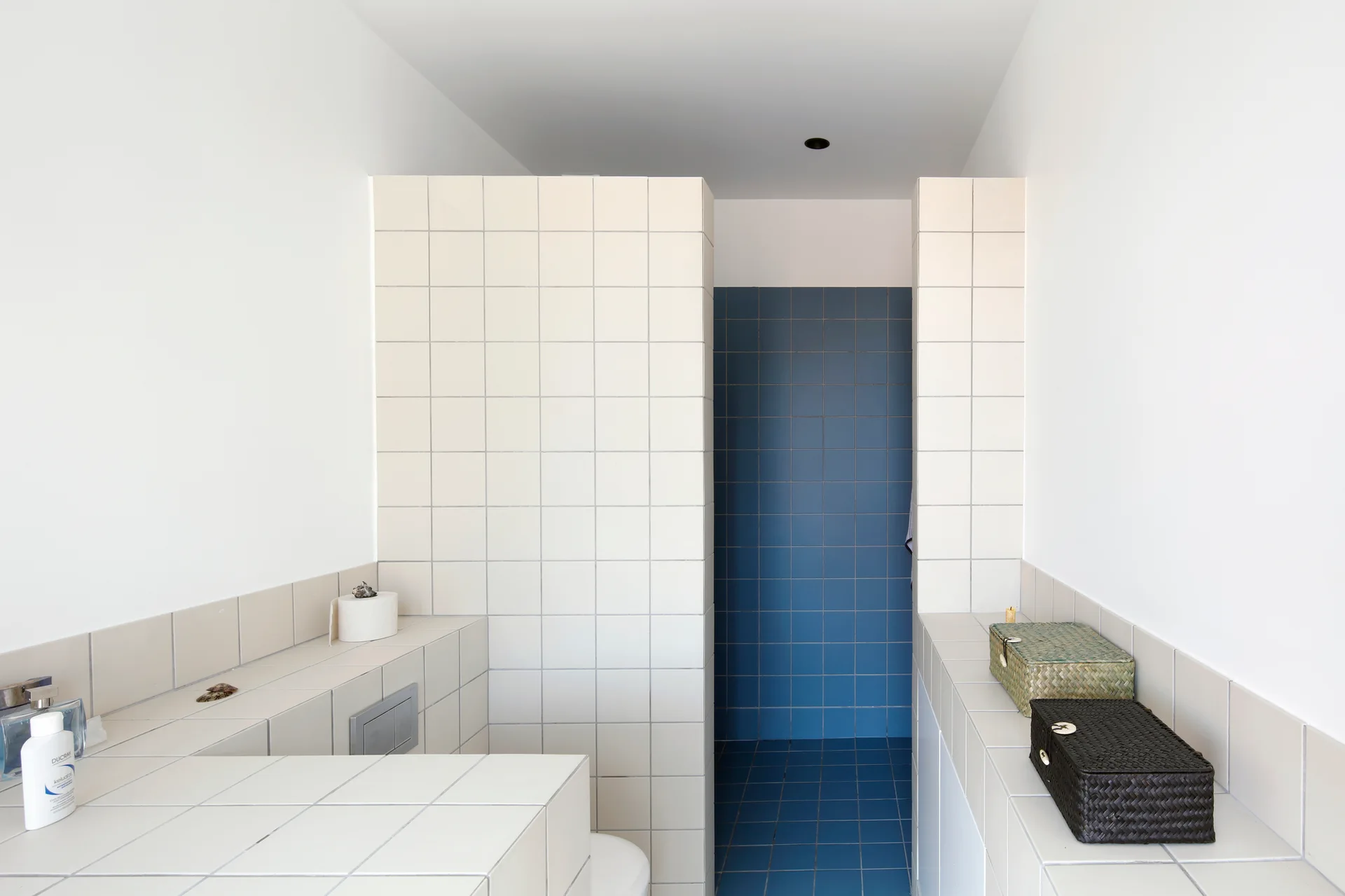 A minimalist bathroom features off-white square grid tiled partition walls and low ledges next to a recessed shower lined with deep blue square tiles.
