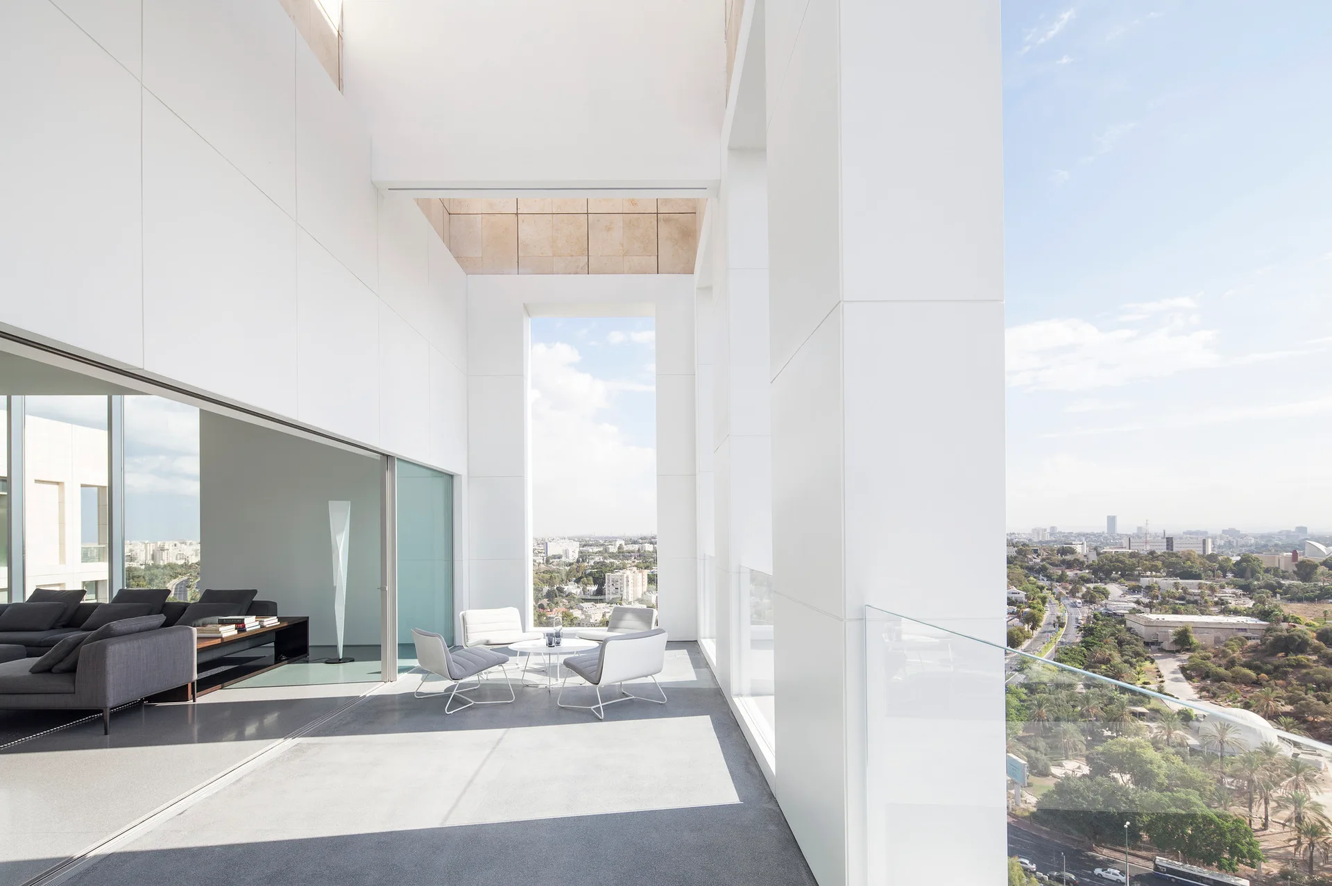 A high-rise terrace with white structural columns, grey outdoor lounge furniture, and a glass railing overlooking a city skyline.