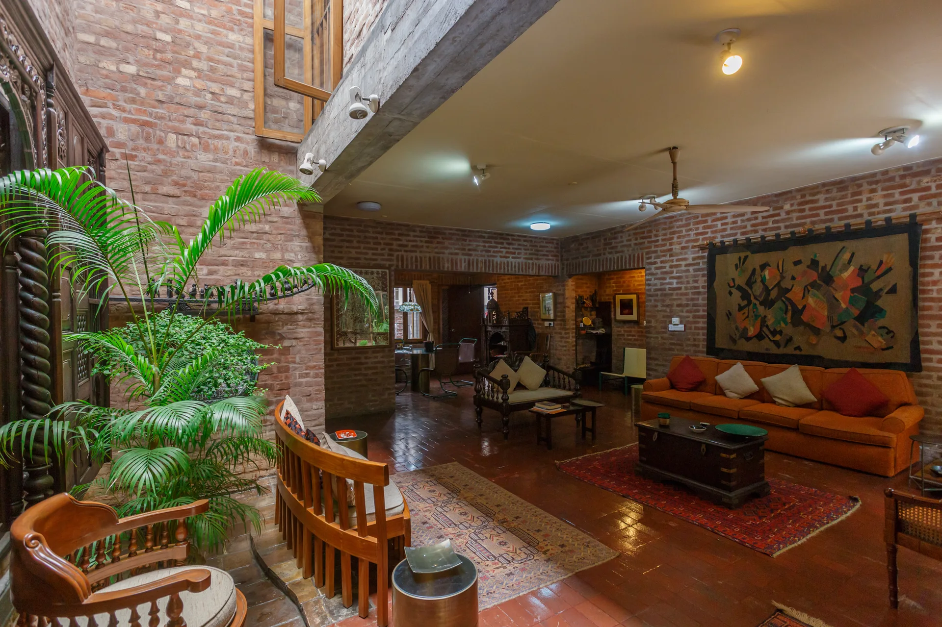 A spacious living room features exposed brick walls, reddish terracotta tile flooring, an orange sofa, and curved wooden seating under a concrete beam.