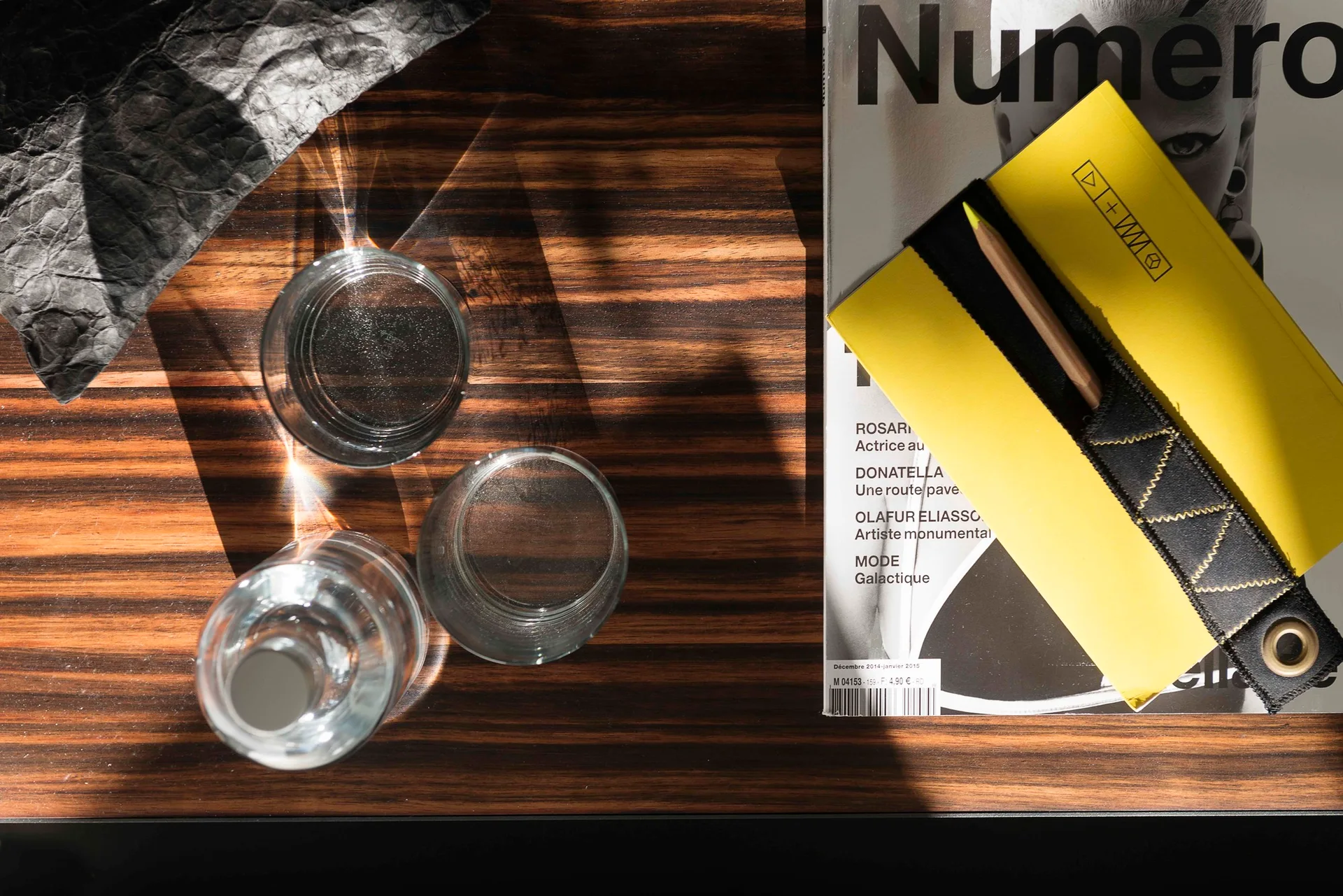 A top-down view of a striped wood table styled with two clear glasses, a magazine, and a bright yellow notebook.