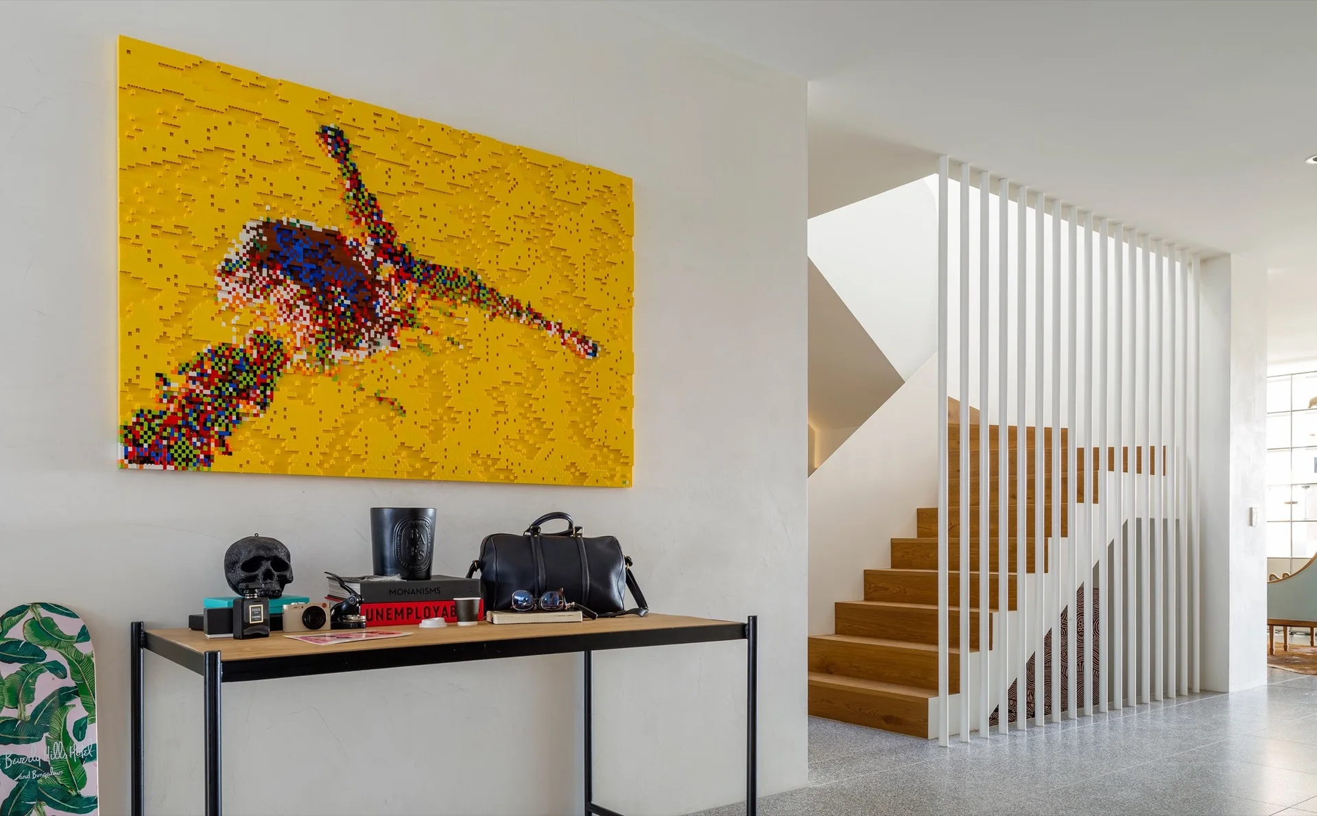 A modern hallway features a black console table under a large yellow artwork next to a staircase behind white vertical slats.