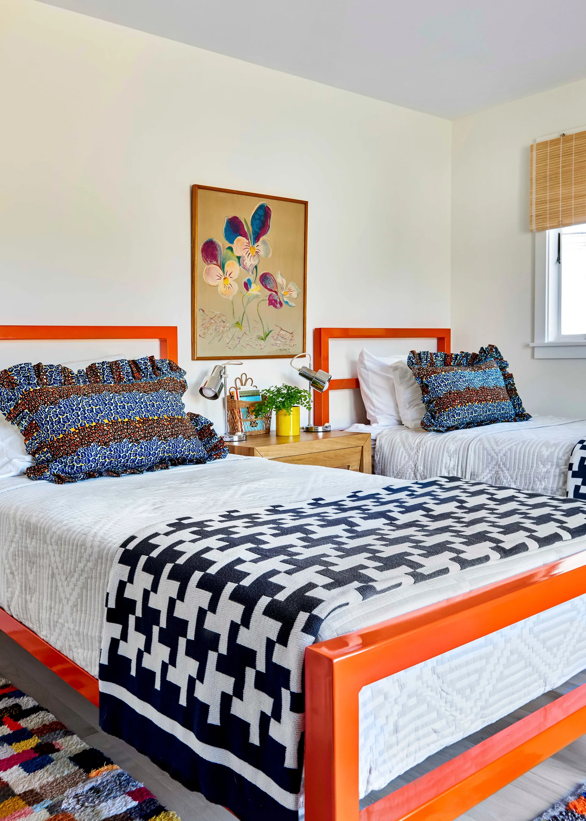 A bright bedroom with two twin beds featuring glossy orange frames, patterned throw pillows, a houndstooth blanket, and a wooden nightstand.
