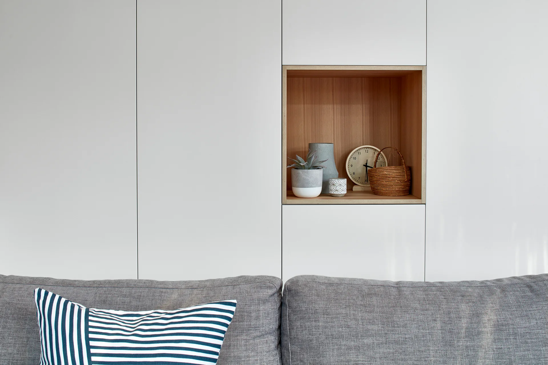 A gray fabric sofa sits below a flat-front white cabinet wall featuring a recessed wood niche styled with a clock, small basket, and plants.