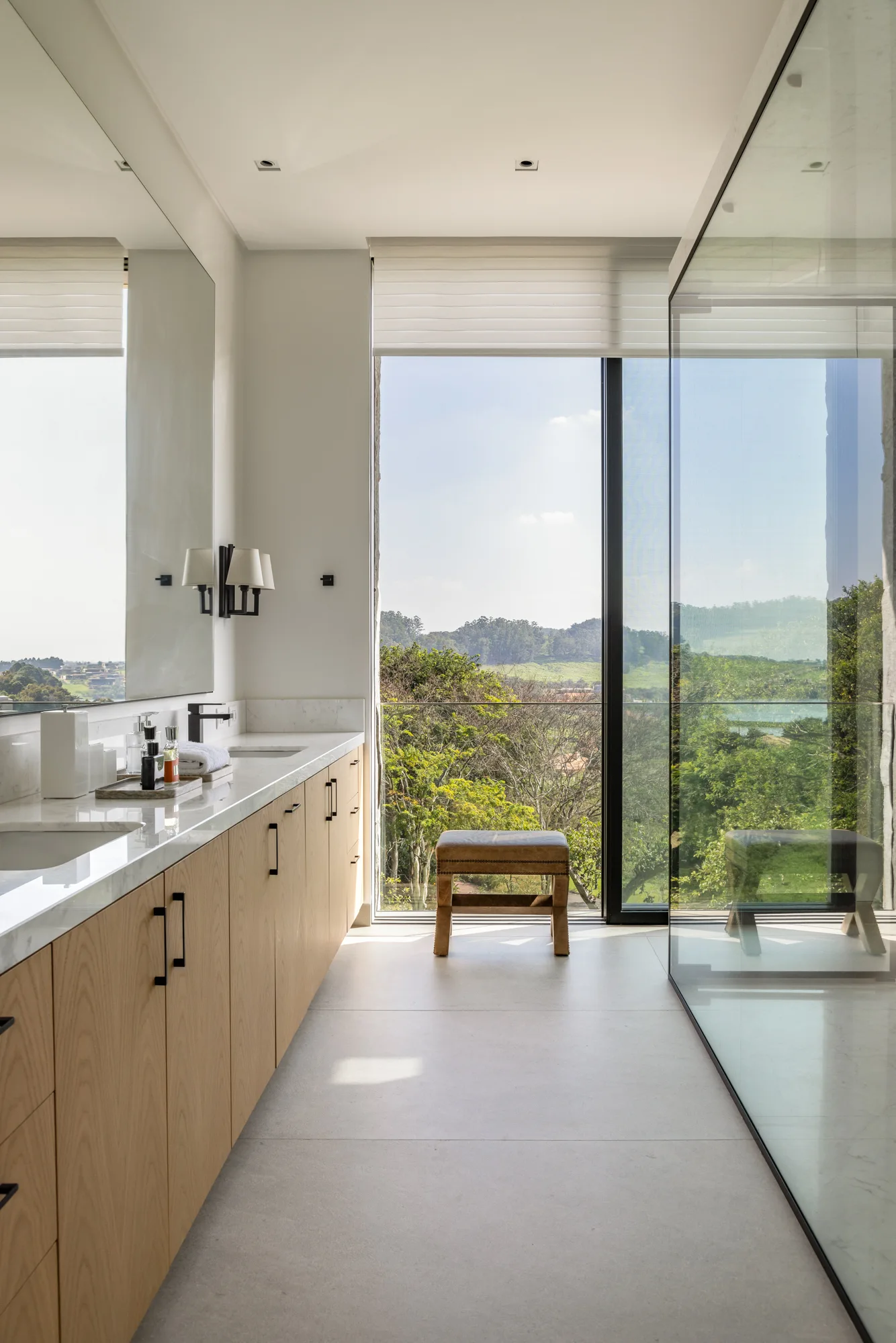 A minimalist bathroom features a light wood vanity with a stone countertop, a large wall mirror, and a floor-to-ceiling window overlooking a landscape.