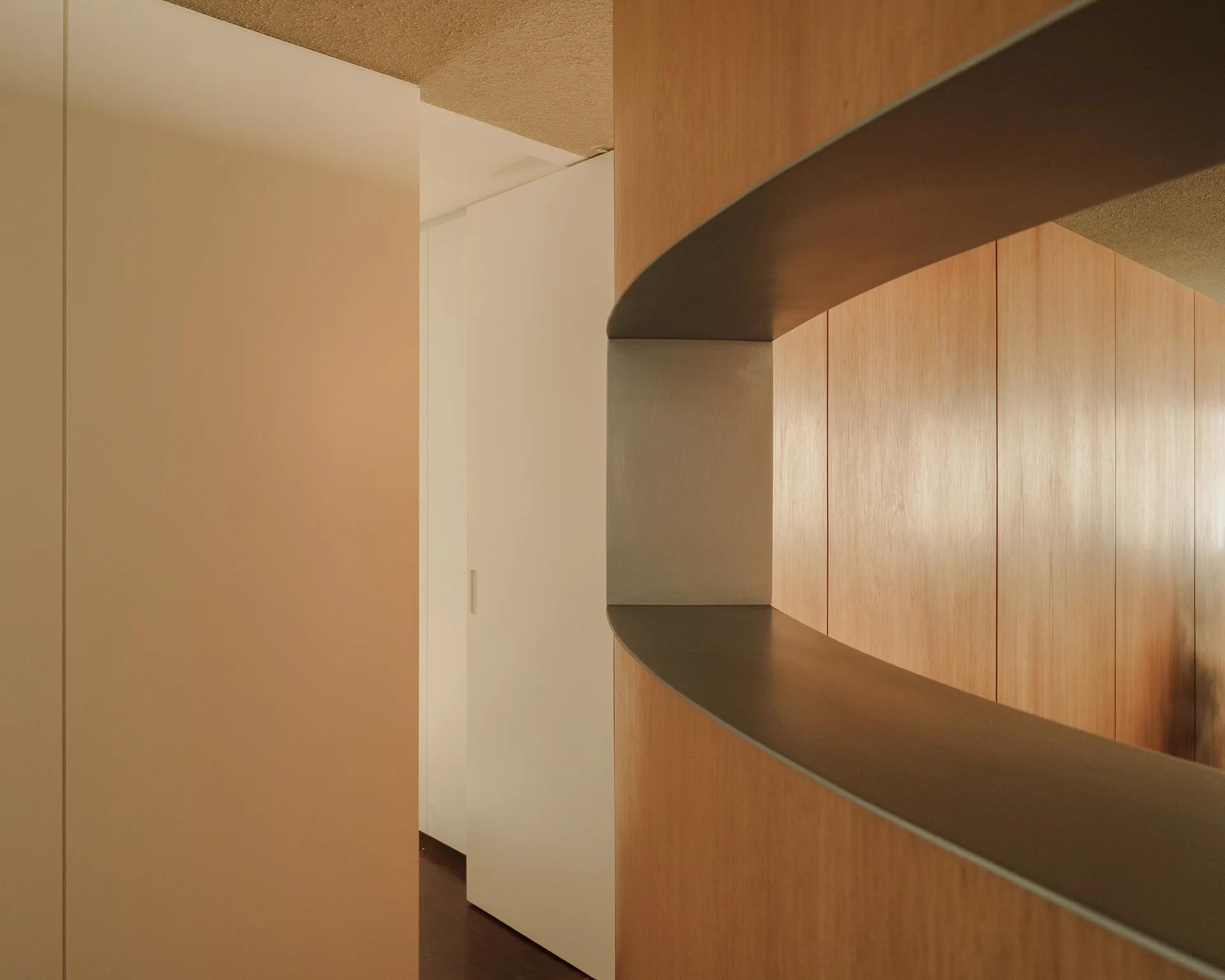 A minimalist hallway corner shows a curved light wood wall with a recessed dark grey shelf next to a flush white door.