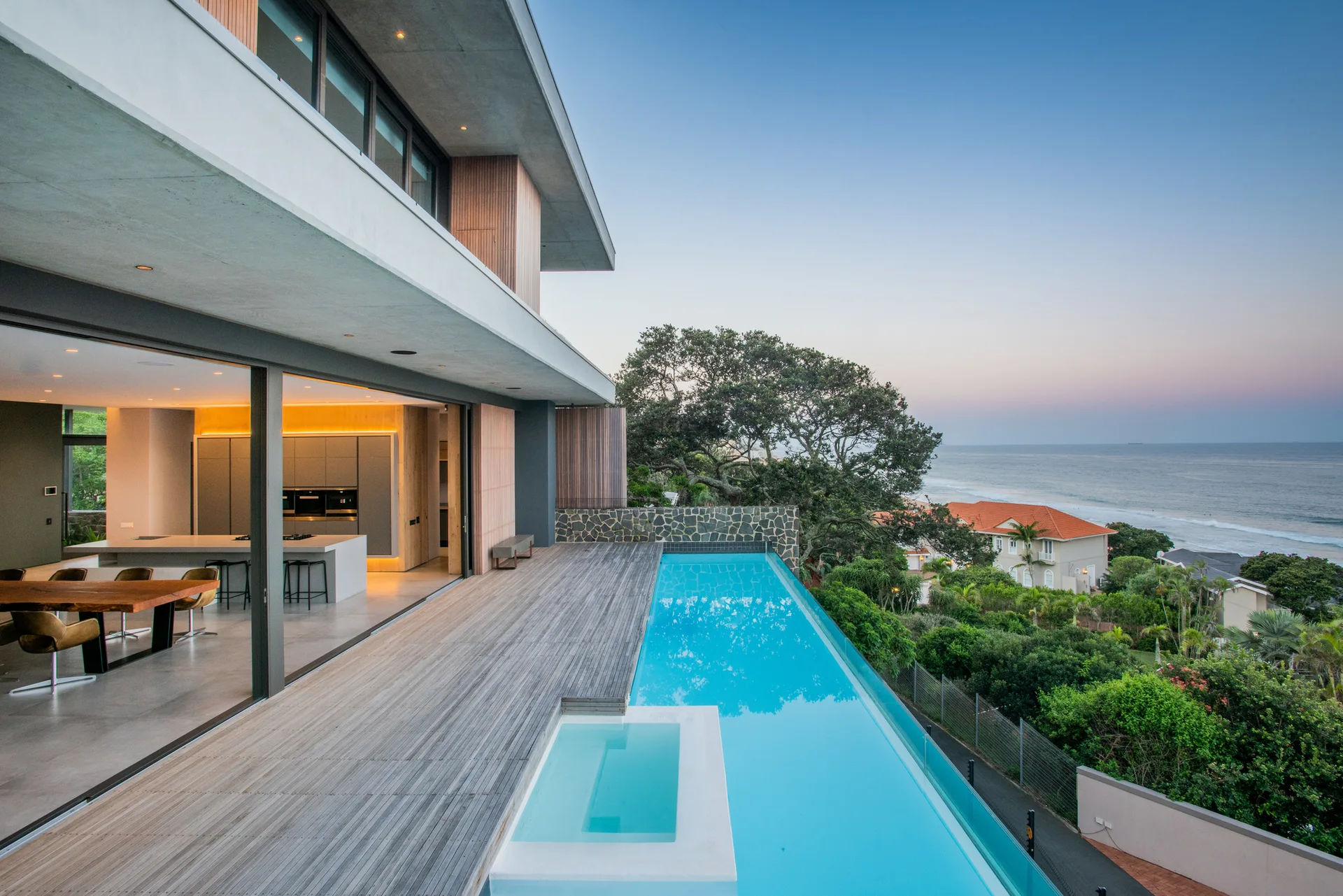 A modern wood deck features a long blue infinity pool overlooking the ocean, adjacent to an open-plan kitchen and dining area.