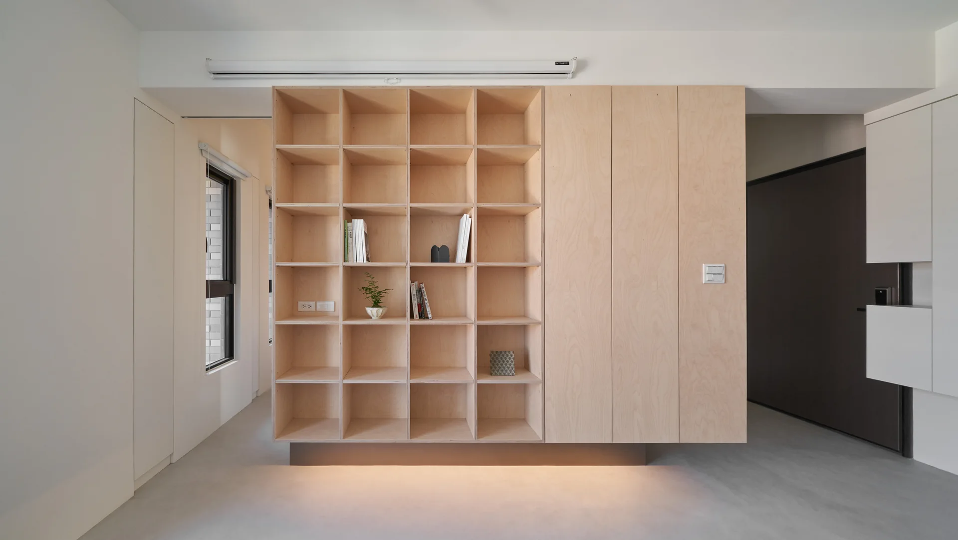 A minimalist plywood shelving unit with open square compartments, flat-panel cabinet doors, and warm lighting glowing from under the recessed base.