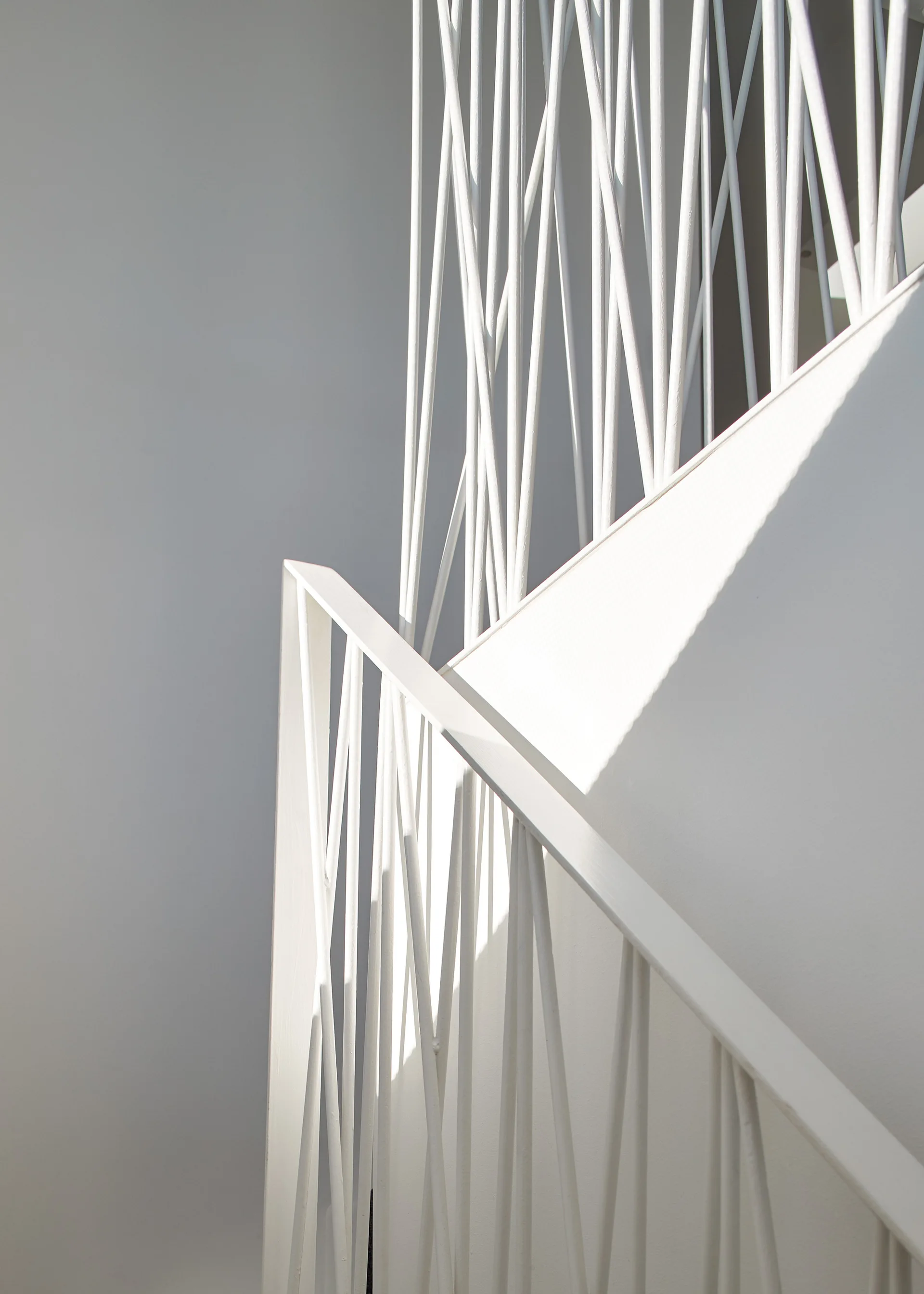 A close-up of a white metal staircase railing with angular, intersecting spindles casting sharp geometric shadows against a matte white wall.