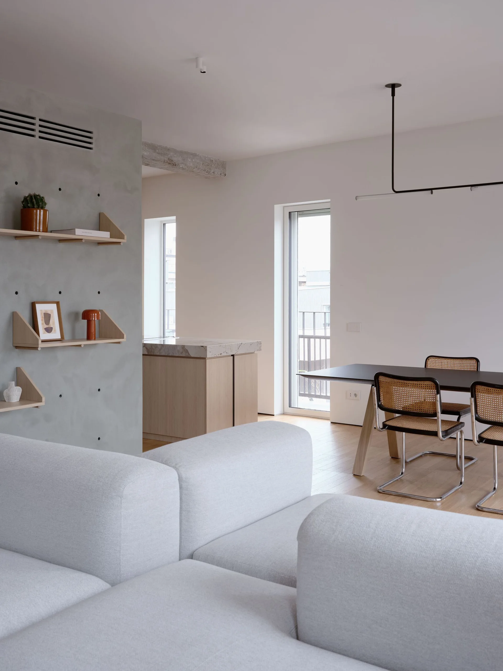 An open-concept living area features a light grey modular sofa, a concrete accent wall with wooden shelves, and cane-back chairs around a dining table.