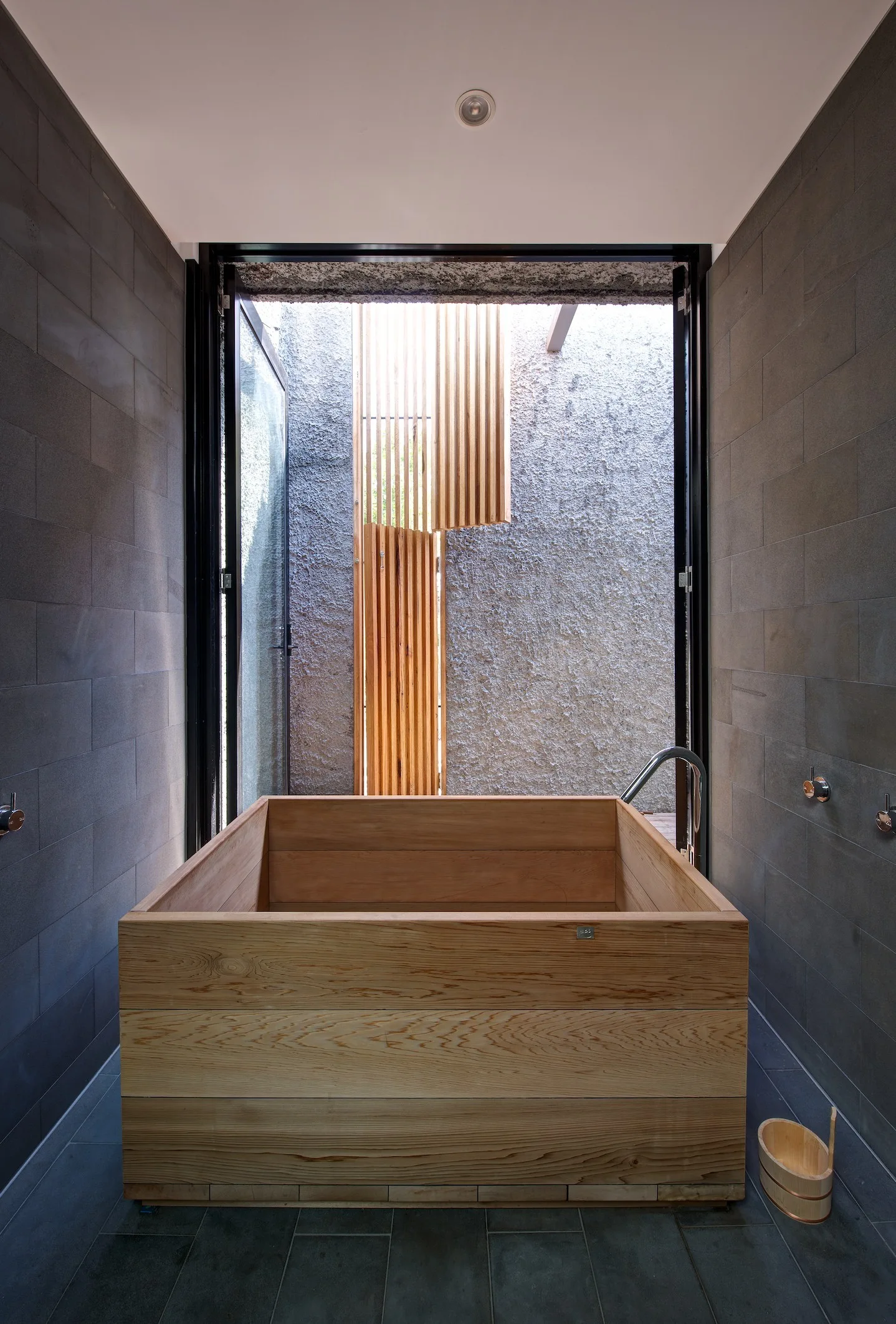 A square wooden soaking tub sits between dark grey tiled walls with an open doorway leading to an outdoor wooden slatted privacy screen.