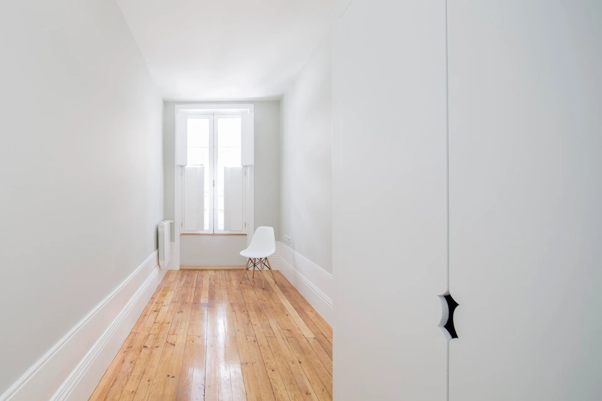 A long, narrow white hallway features polished wood plank flooring, tall baseboards, and a single white chair near a window at the end.