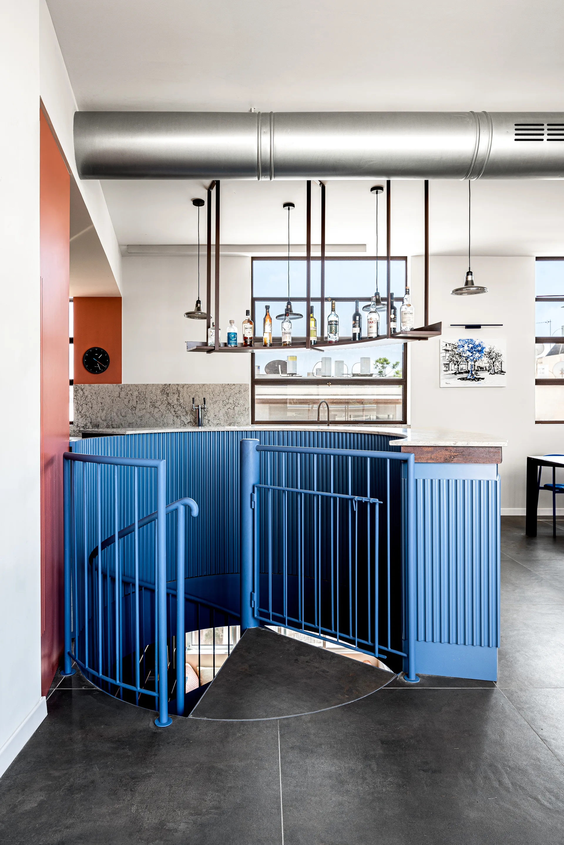 A blue spiral staircase with a fluted metal railing is integrated into a bar area featuring a suspended bottle rack and exposed industrial ductwork.