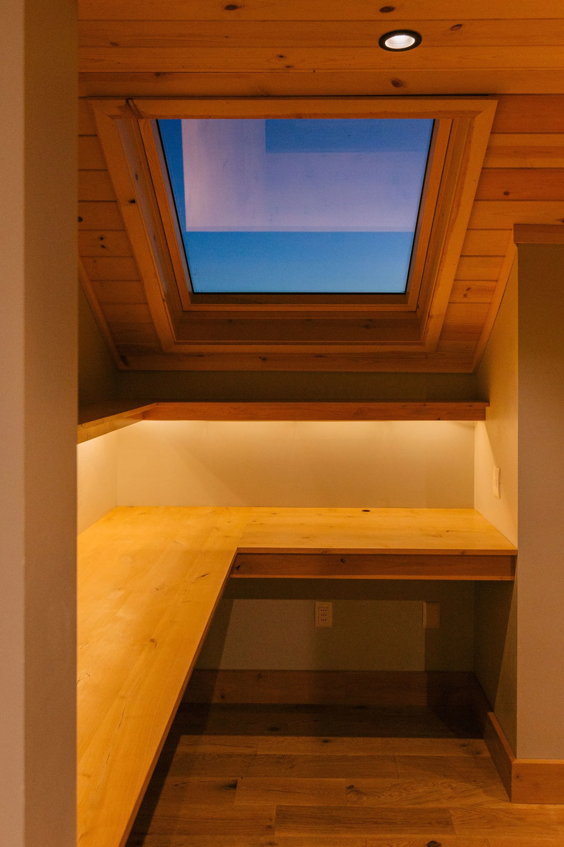 A small built-in wooden L-shaped desk and floating shelf sit within a cozy office nook beneath a sloped wood plank ceiling with a skylight.