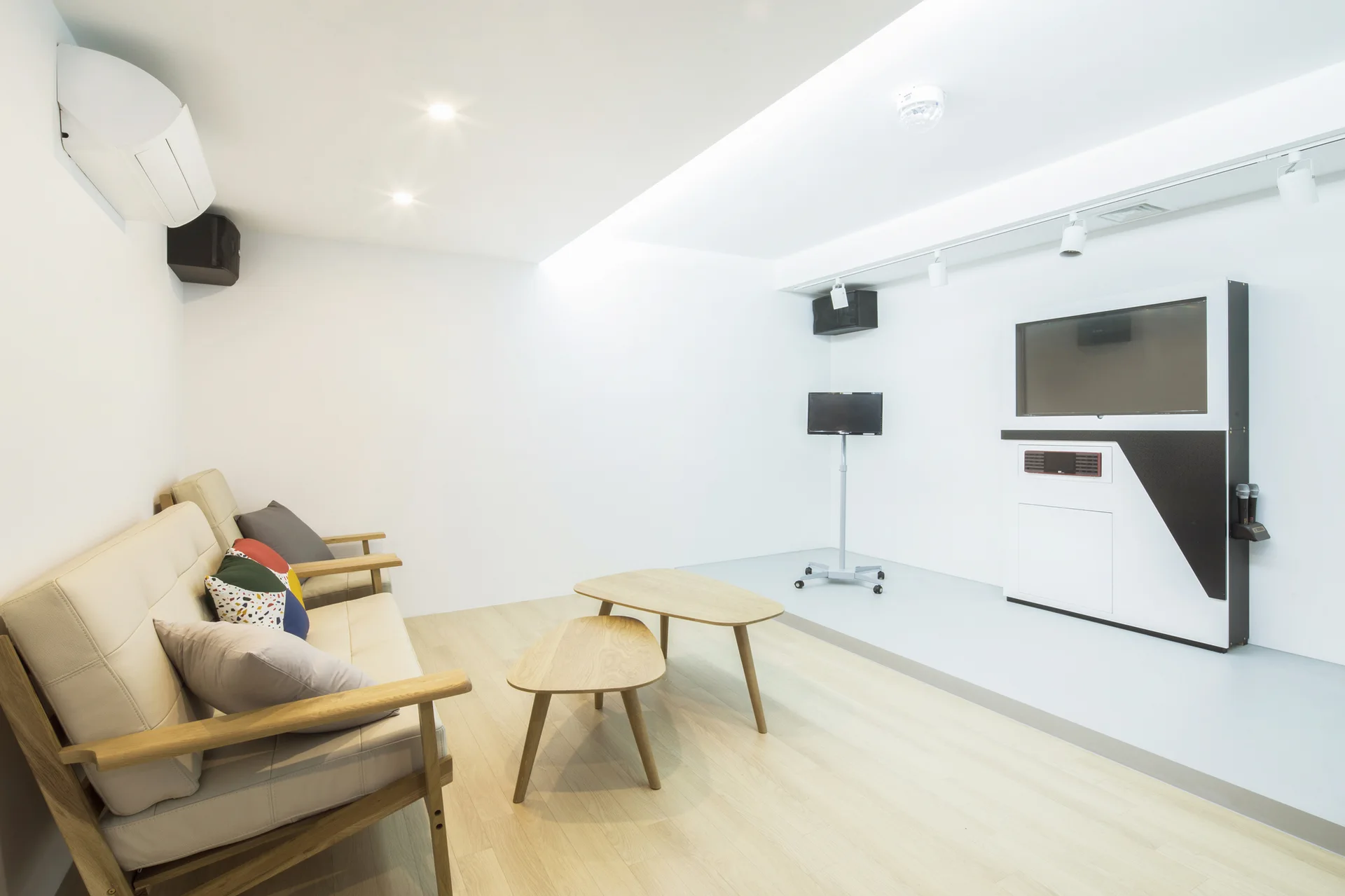 A minimalist media room features a light wood-framed beige sofa, two triangular wooden coffee tables, and a white built-in television unit.